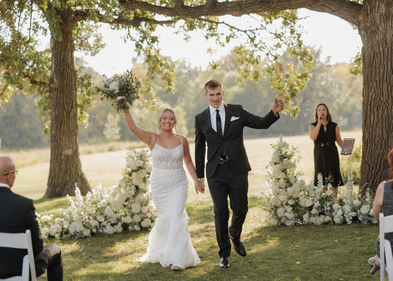 Newlyweds celebrate with raised arms during outdoor ceremony with floral arrangements under oak trees at sunset.