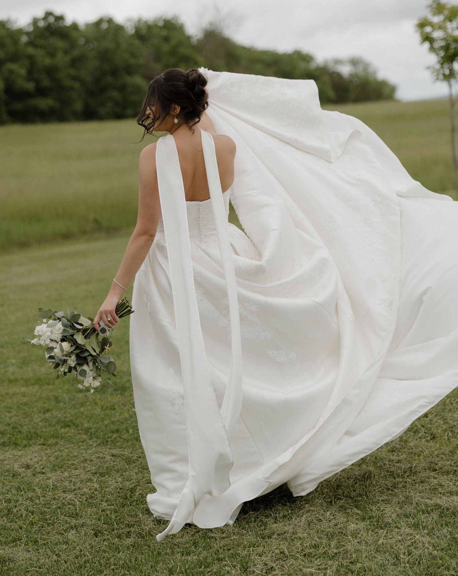 Bride in flowing white wedding gown with dramatic cape veil, holding bouquet, standing on lush green grass.