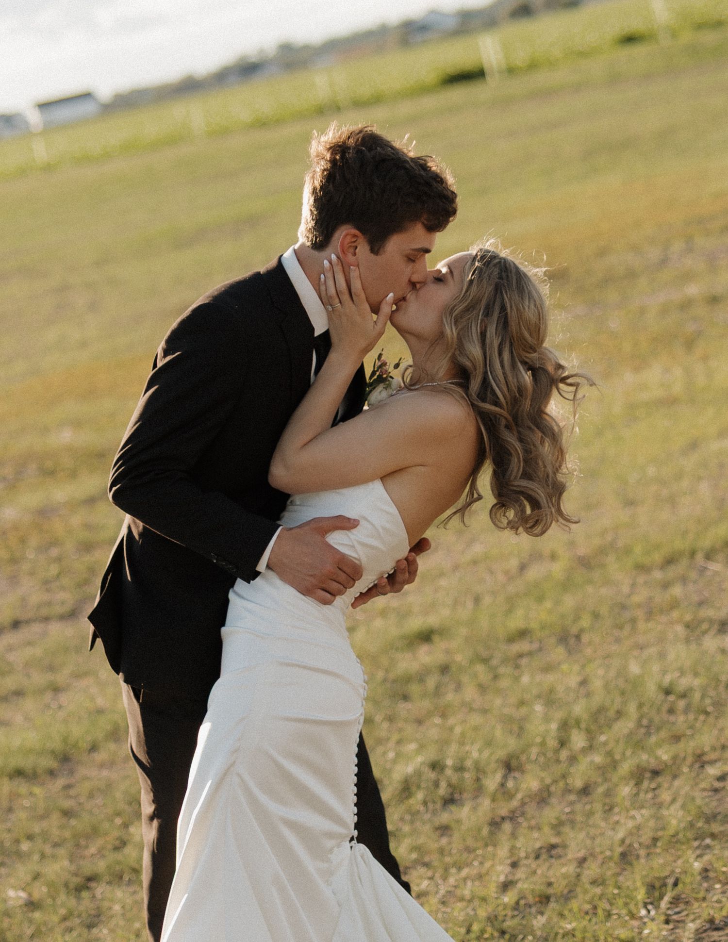 A bride and groom share a romantic kiss in an open field, the bride in a white gown and groom in a black suit.