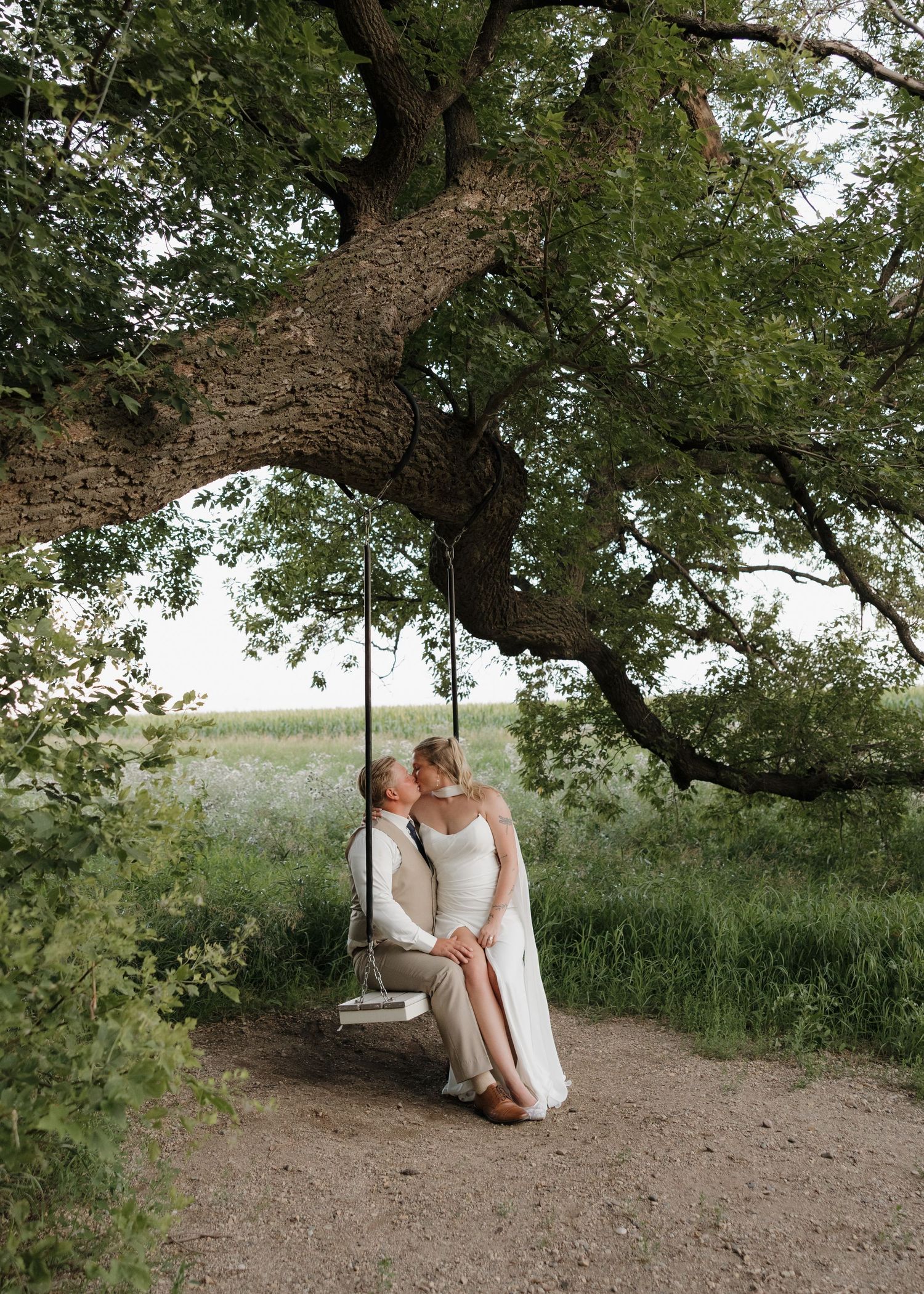 A couple sits together on a rustic wooden swing beneath a sprawling tree canopy on a dirt path.