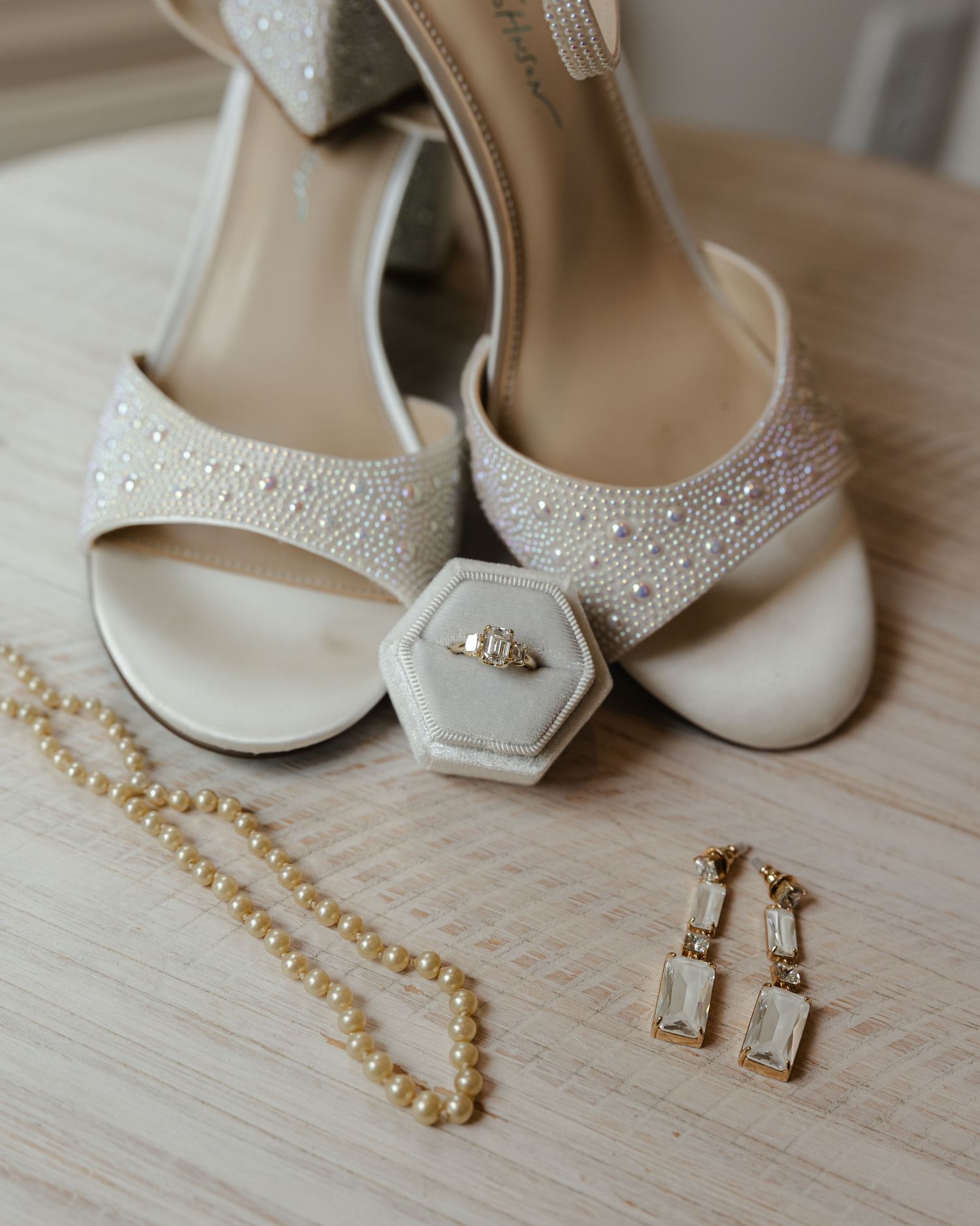 Elegant white bridal shoes with rhinestone details displayed alongside matching gold jewelry and earrings on wooden surface.