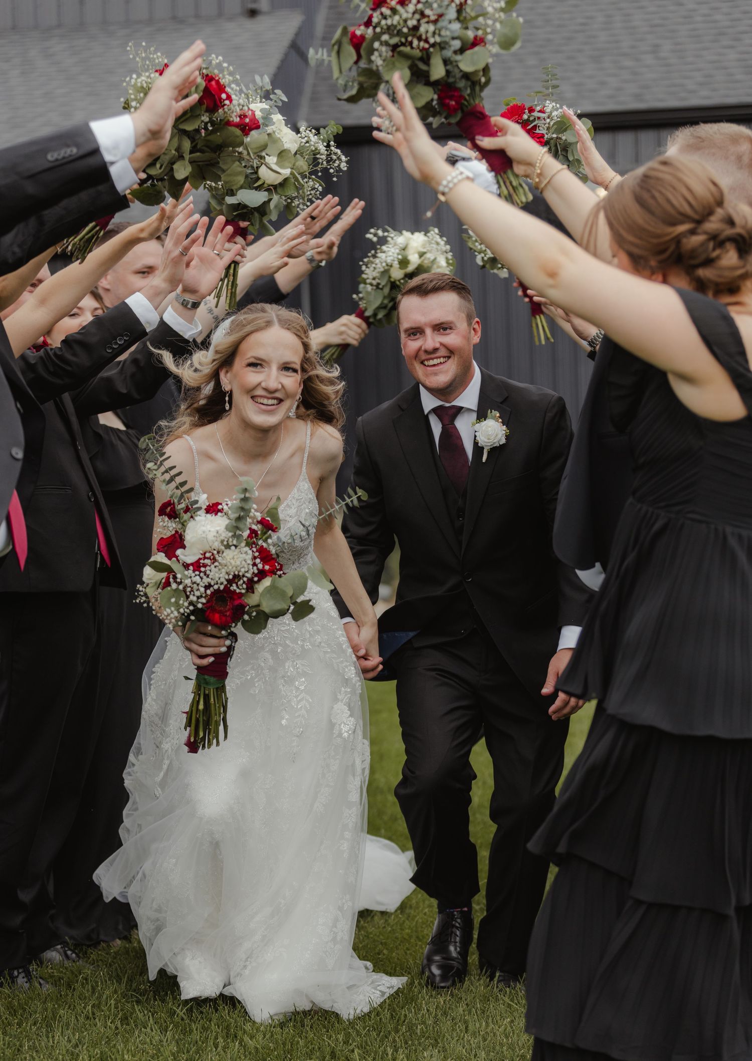 Wedding party celebrates joyfully outdoors with raised arms and bouquets in black and white attire.