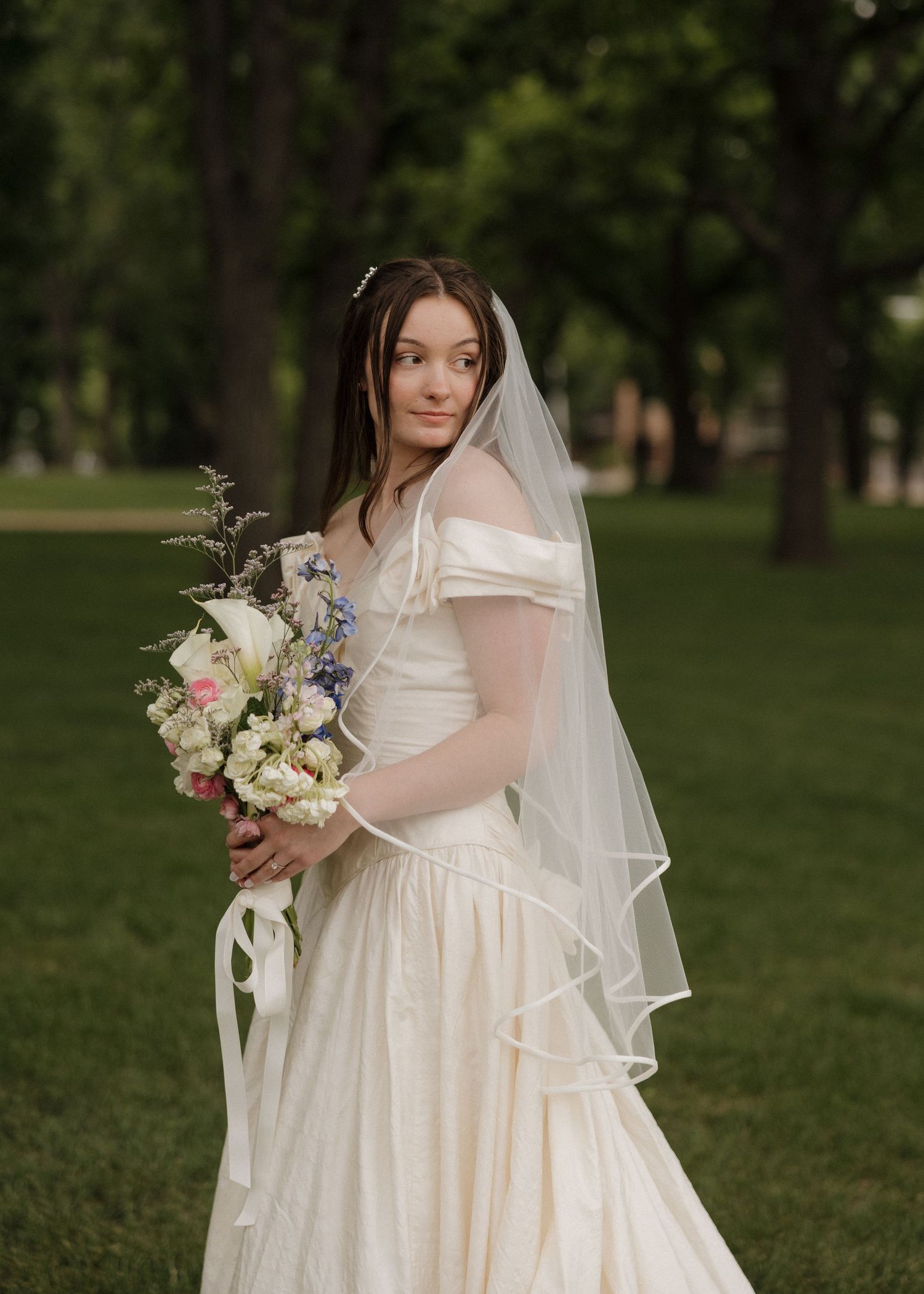Bride in white off-shoulder gown and veil holds floral bouquet outdoors in lush green park setting.