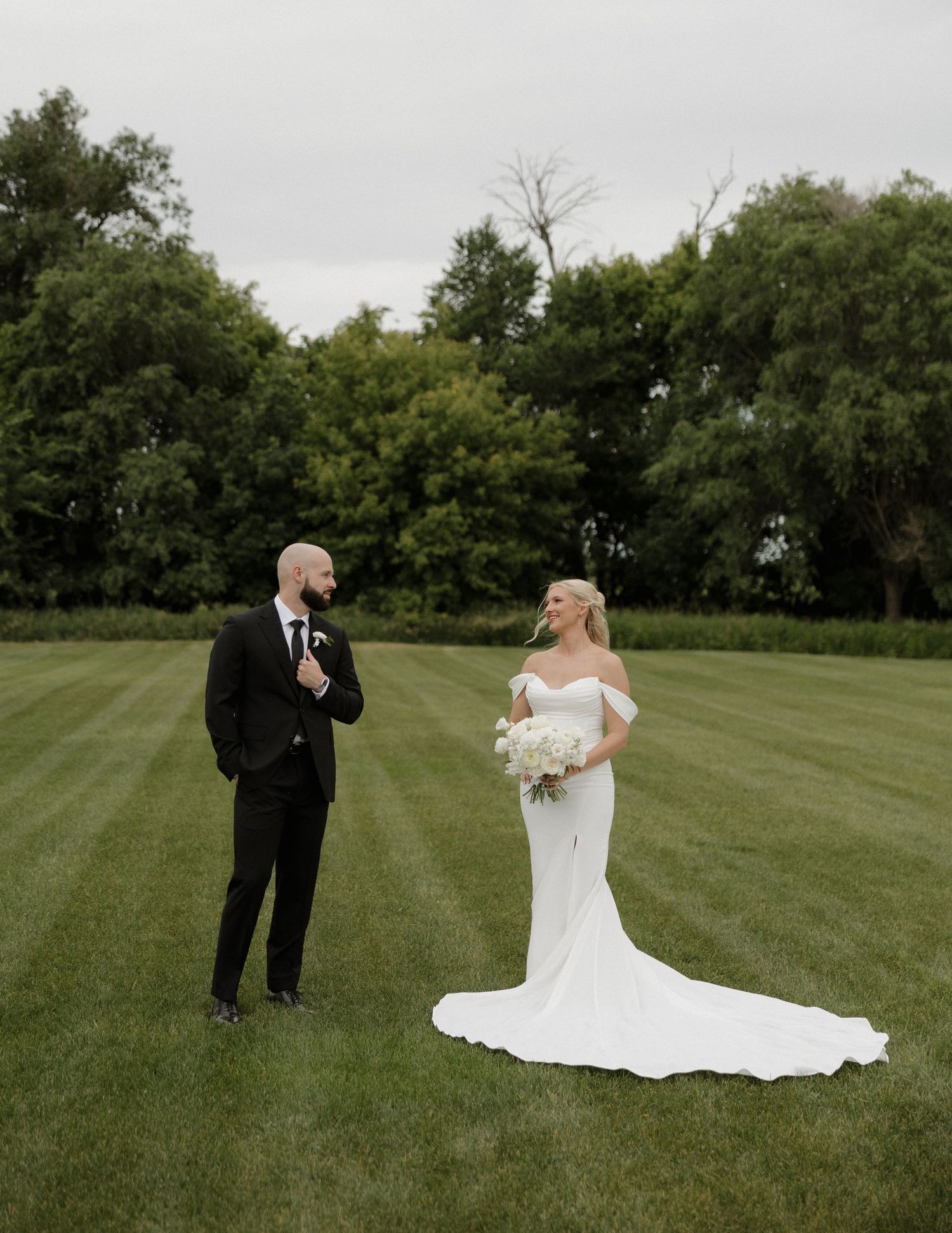 Bride in white off-shoulder gown and groom in black tuxedo share a moment on lush green lawn.