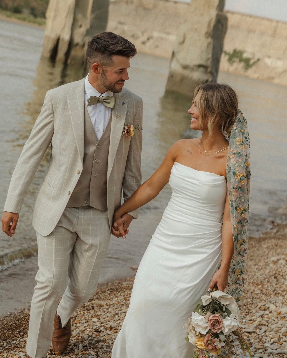 A couple in wedding attire share a romantic moment on a rocky beach, with the bride in white and groom in light beige suit.