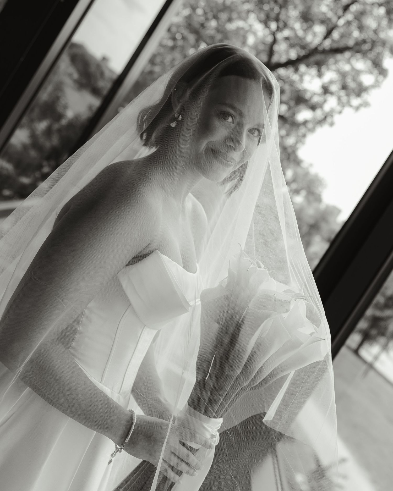 A bride in a flowing white wedding dress and veil poses elegantly in black and white photography.