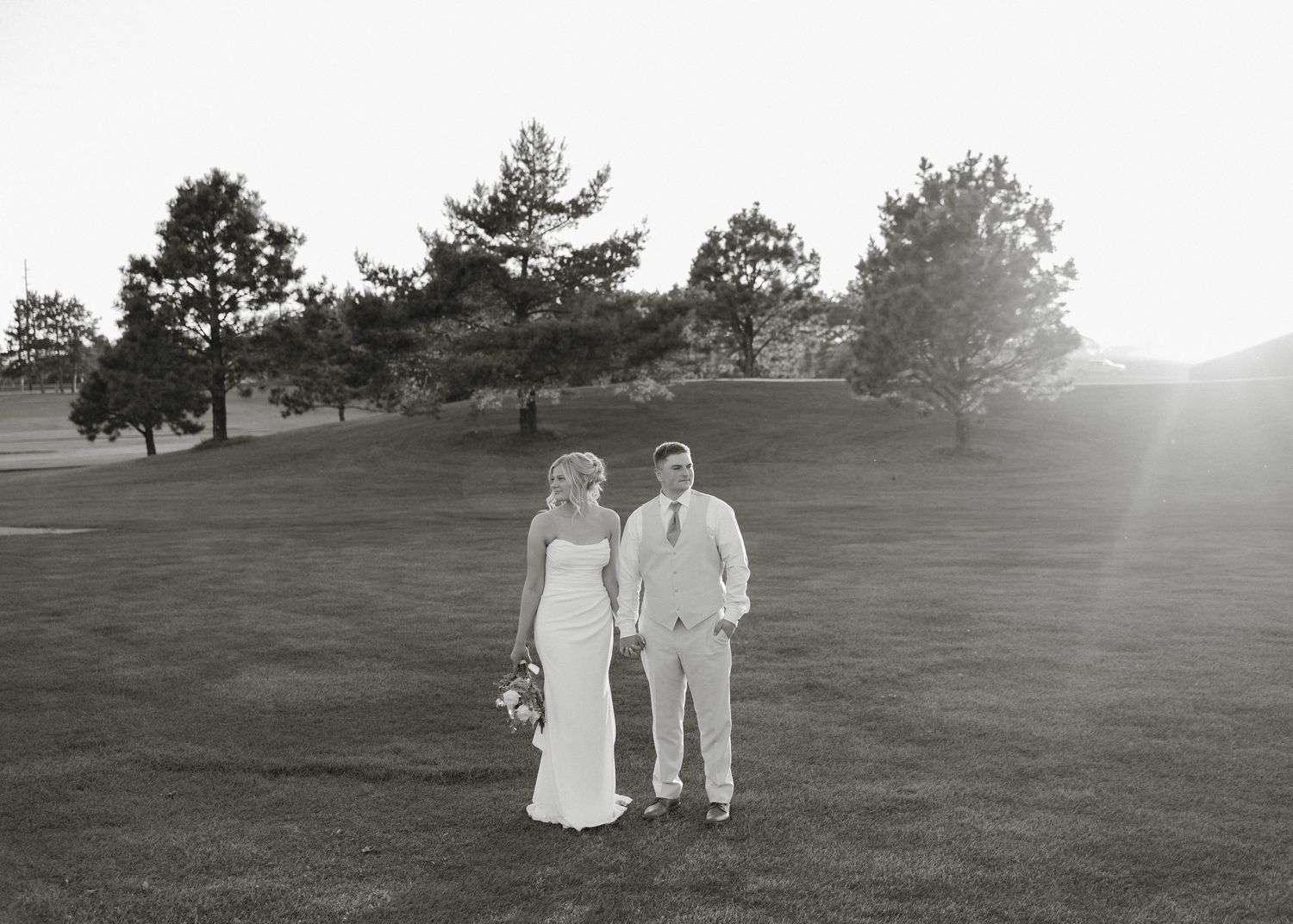 A black and white photo of a couple standing on a golf course with trees in the background at sunset.