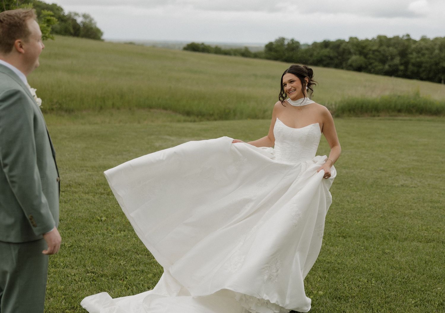 Bride twirling white wedding gown in lush green field while groom in sage suit watches during outdoor ceremony.