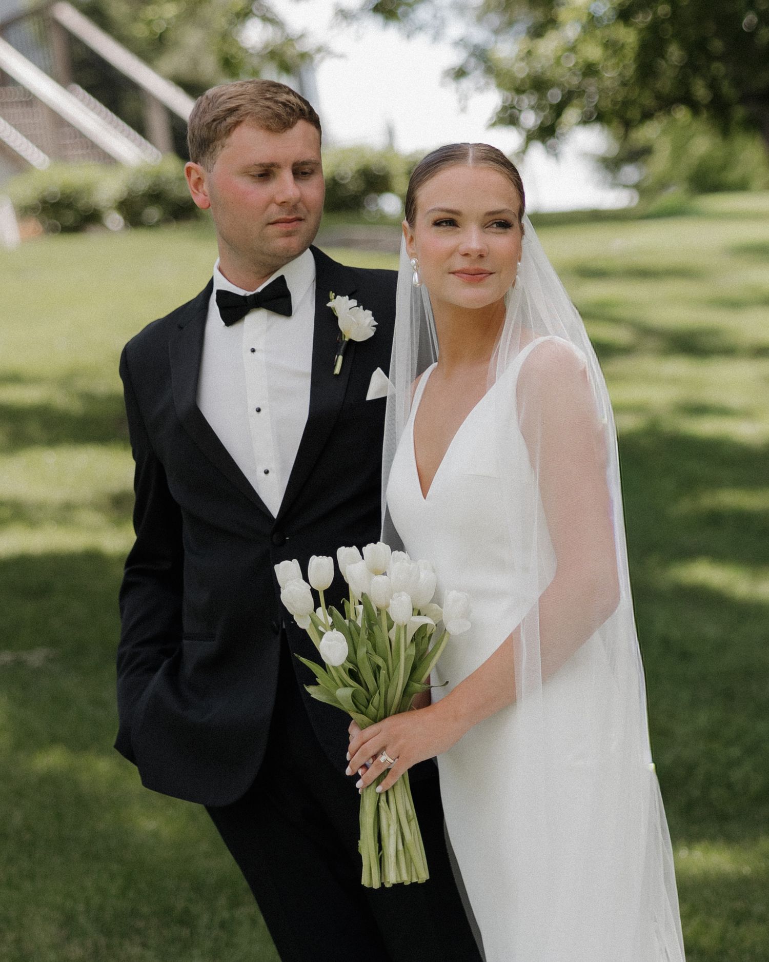 A bride holding white tulips in a sleek gown and veil stands beside a groom in a black tuxedo with bow tie outdoors.