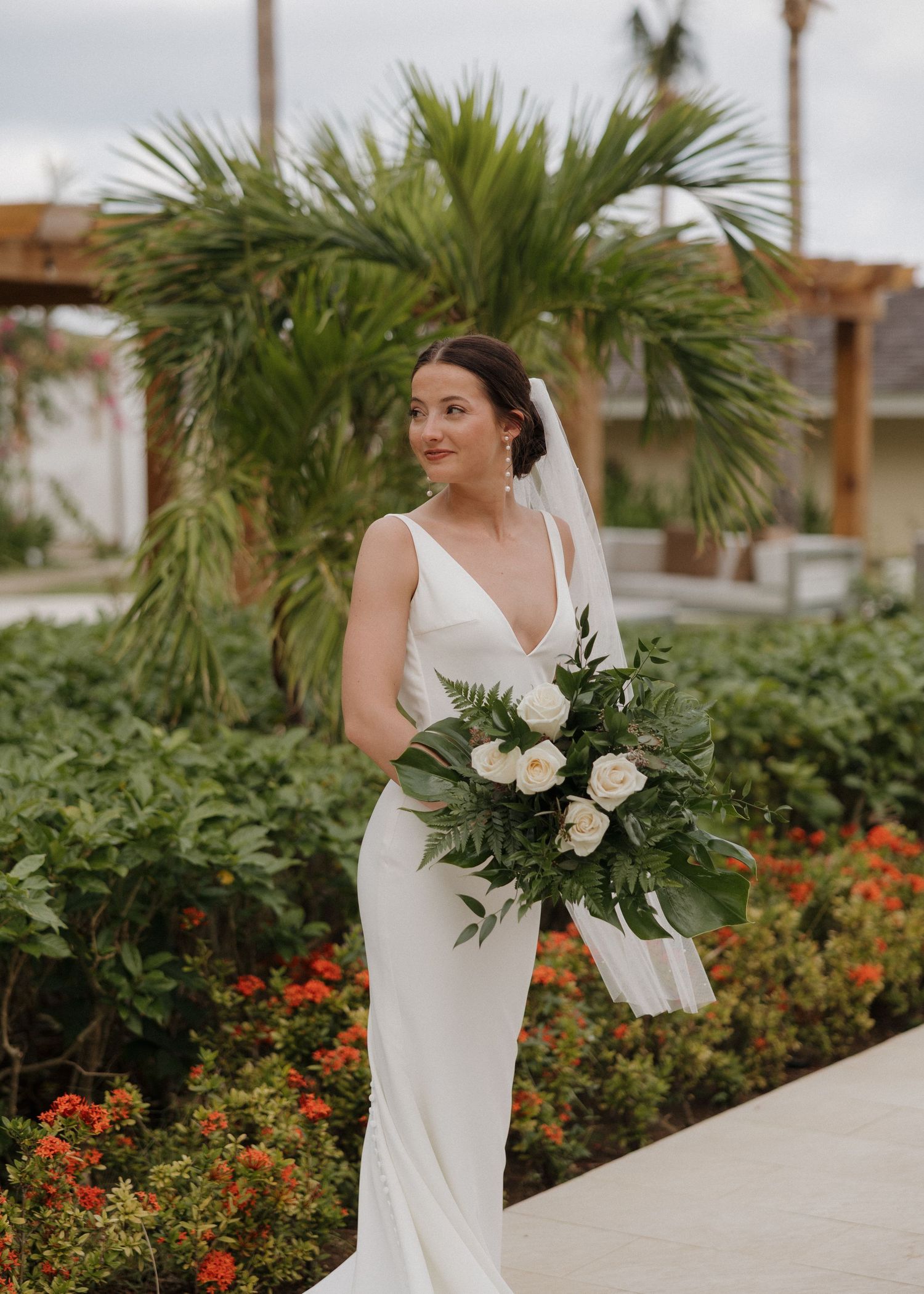 A bride in a sleek white wedding dress poses elegantly among tropical palm trees and vibrant orange flowers.