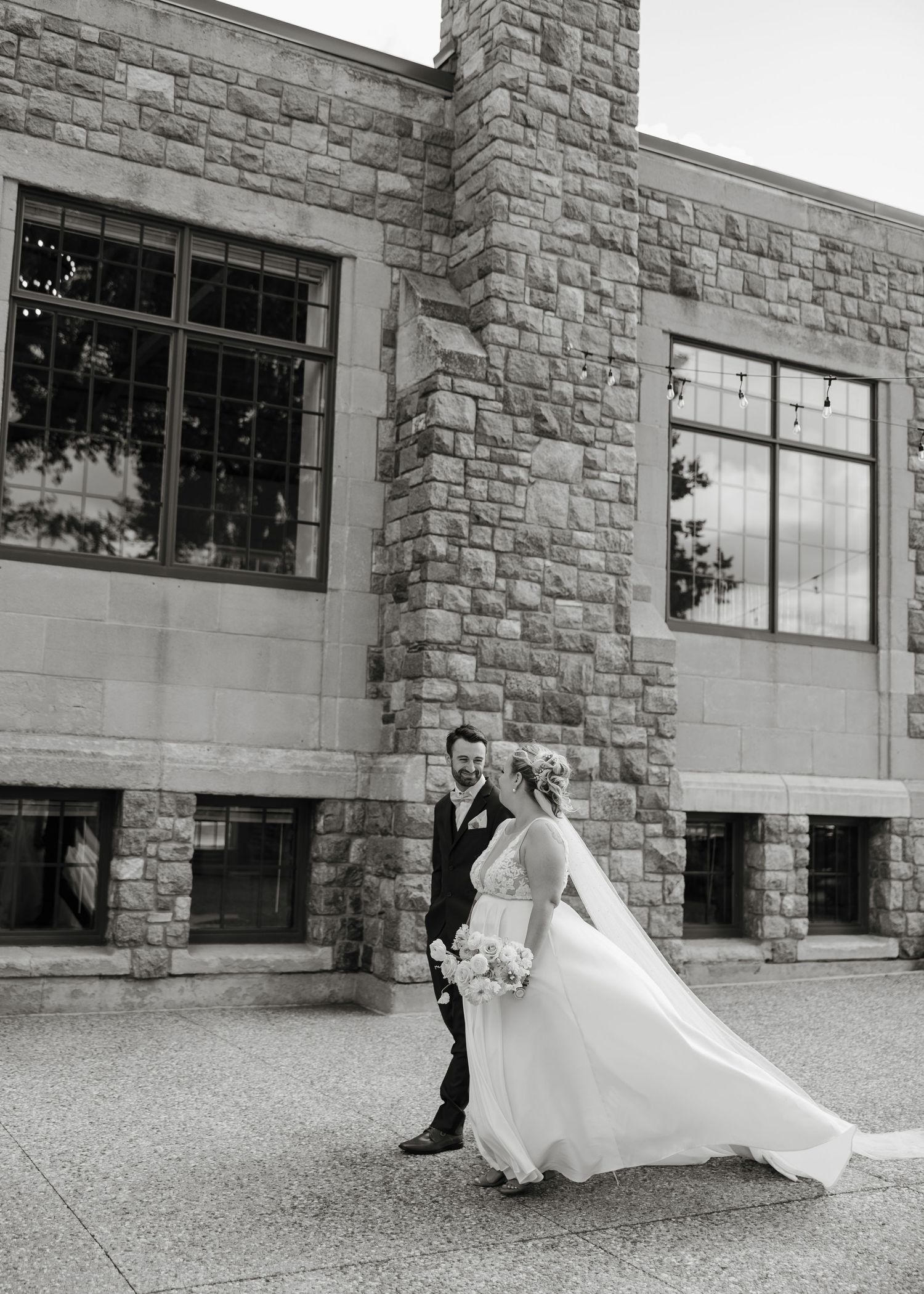 A couple in wedding attire stands outside a stone building with large windows in black and white photography.