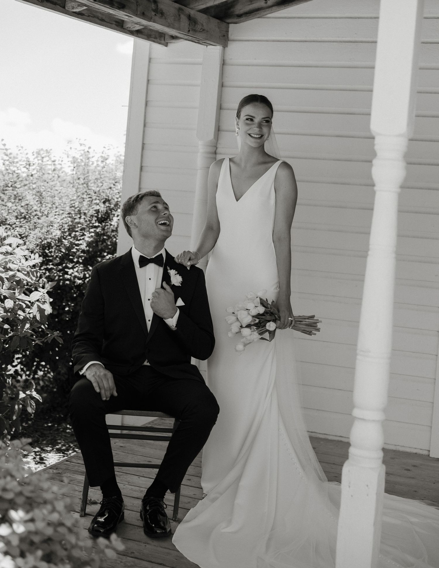 Black and white photo of bride in white gown and groom in tuxedo on a porch, bride holding bouquet.