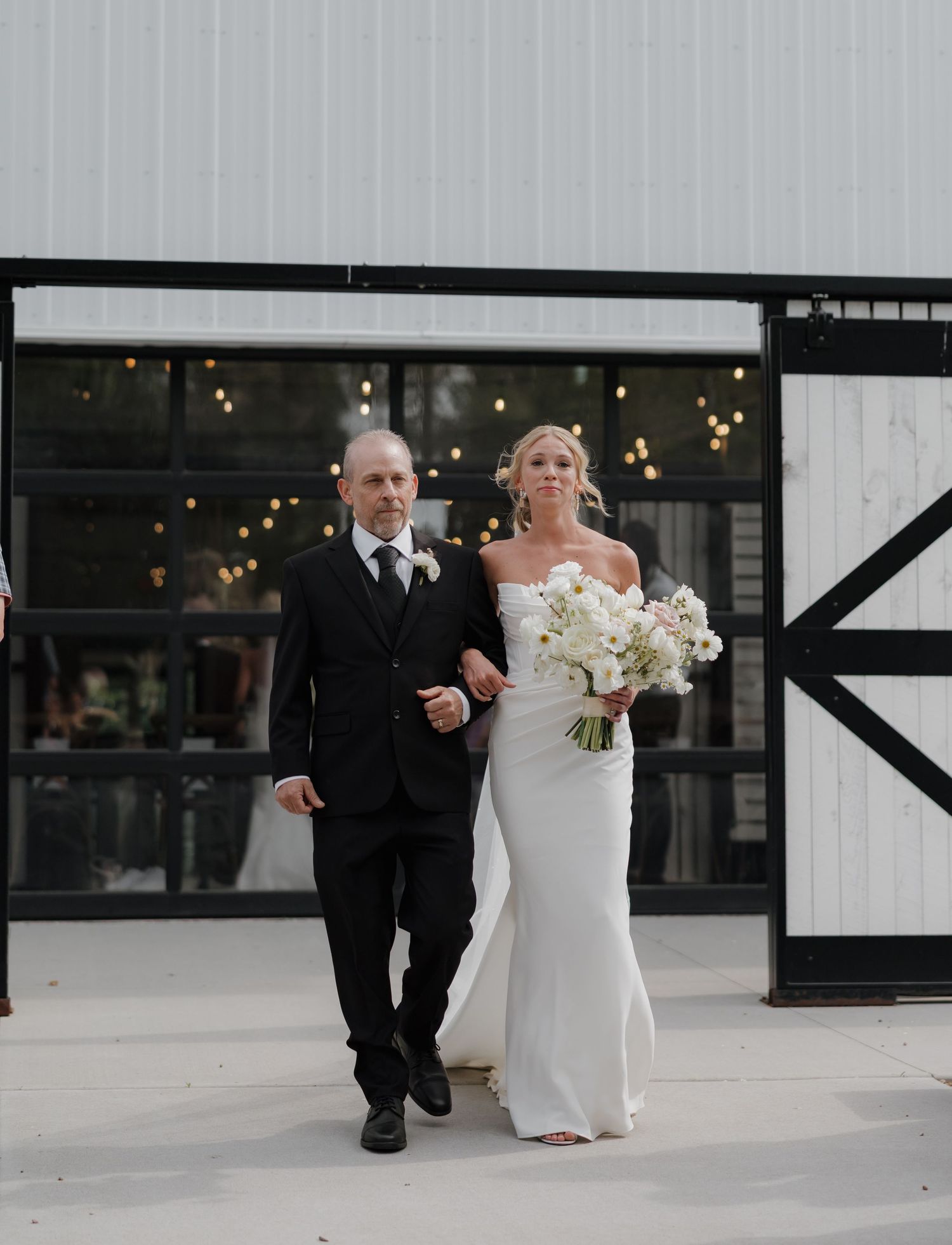 A bride and father walk down a modern white hallway with black accents during a wedding ceremony entrance.