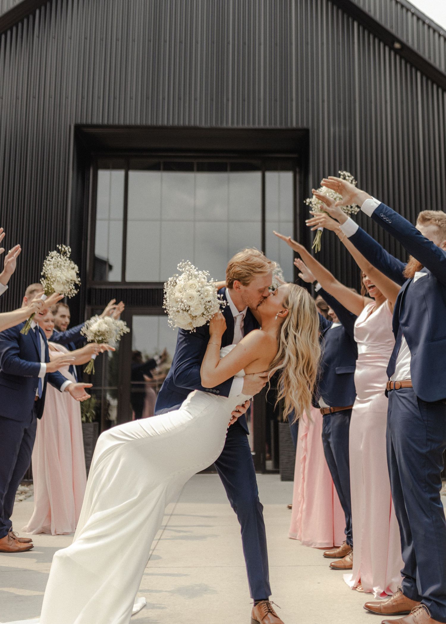 Wedding party celebrates with raised bouquets as couple shares romantic dip kiss in front of modern black barn venue.