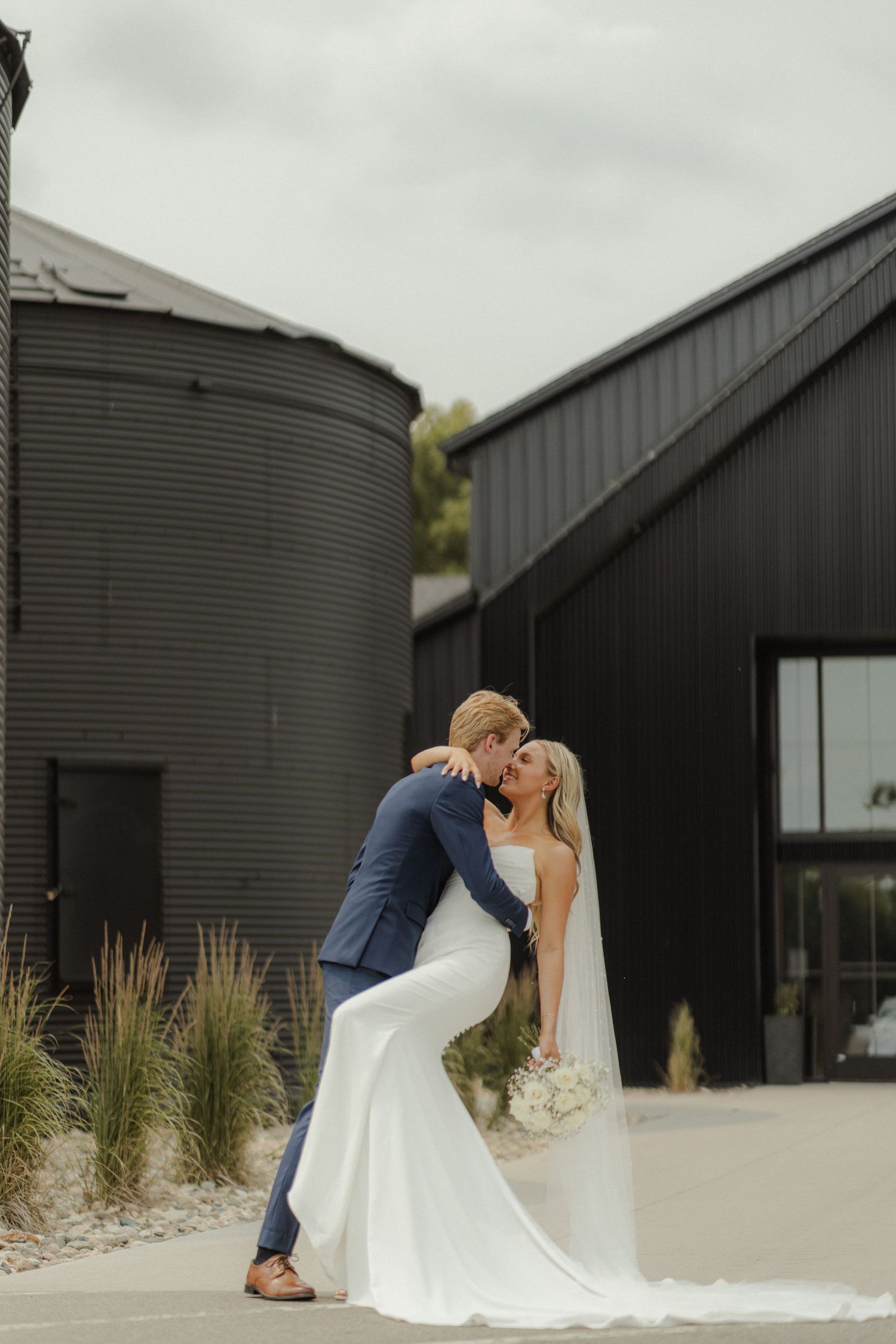A romantic wedding couple shares a tender kiss outside a modern black barn venue.