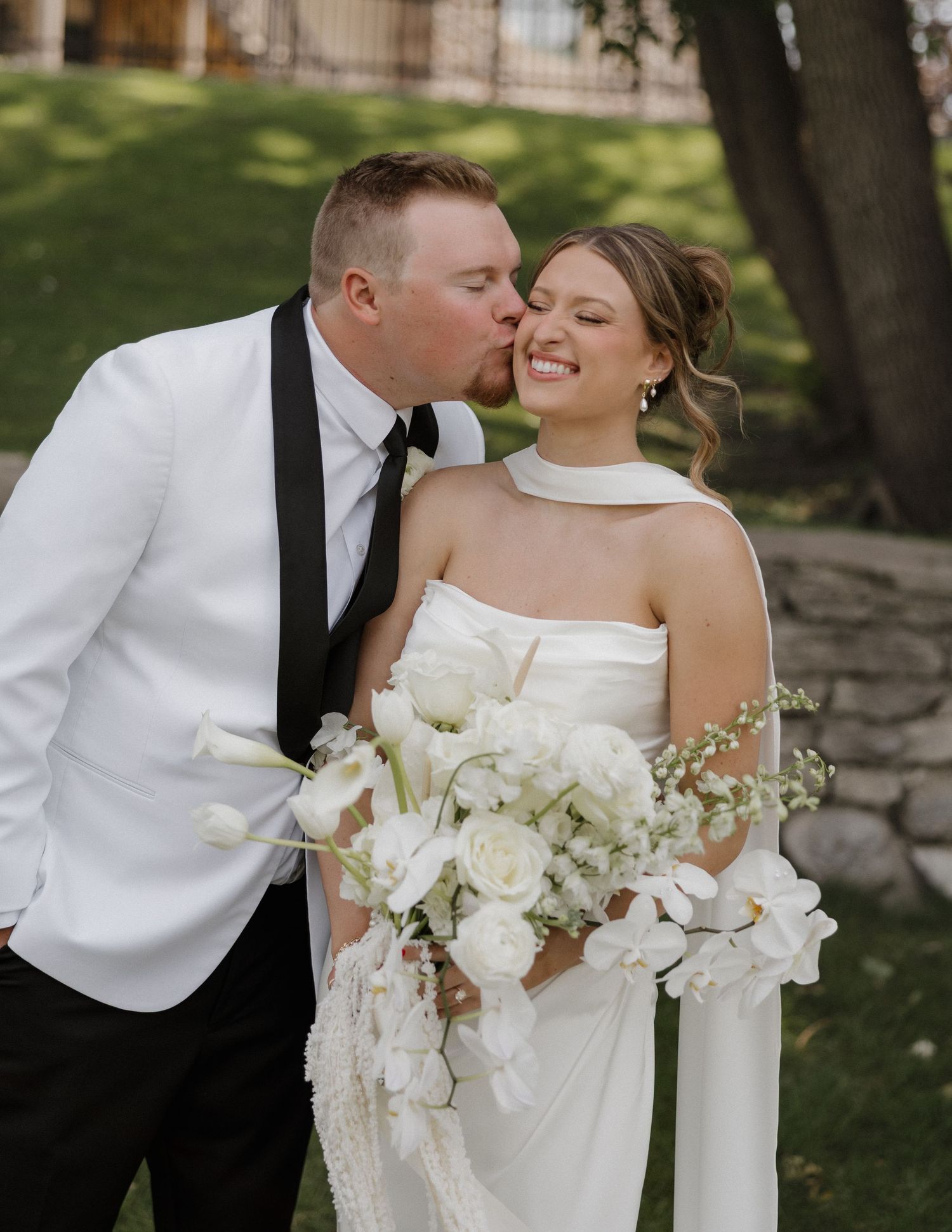 Romantic wedding couple sharing tender moments in garden setting with elegant white floral bouquet.