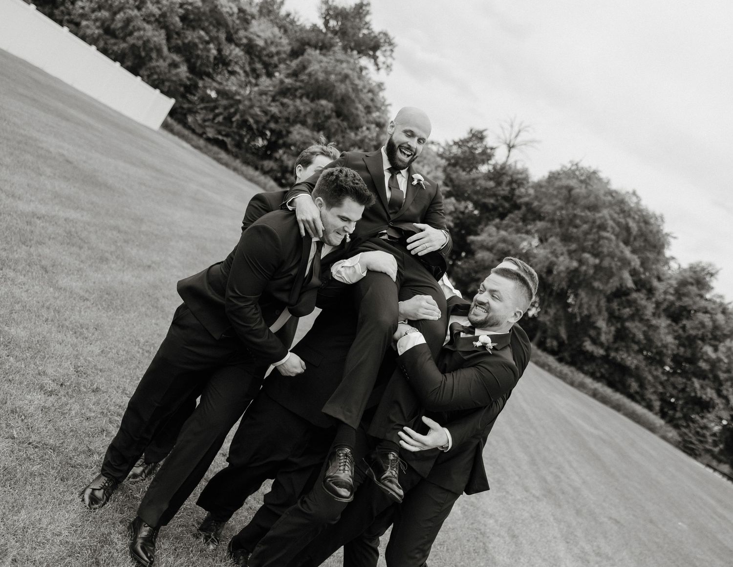 Groomsmen in black suits playfully lifting the groom outdoors on a grassy lawn in a black and white photo.