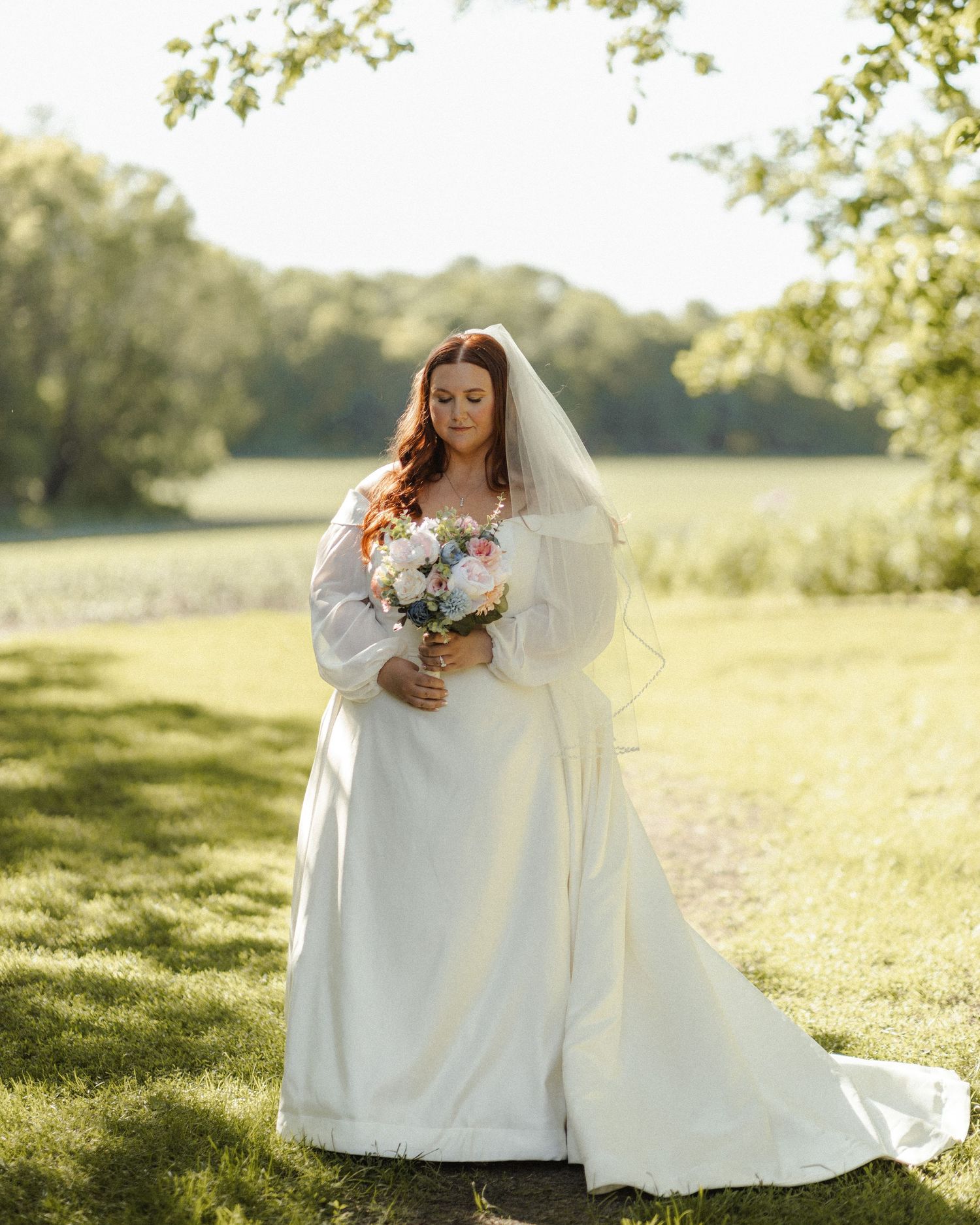 A bride in a flowing white dress sits gracefully on lush green grass during a sunny outdoor wedding photoshoot.
