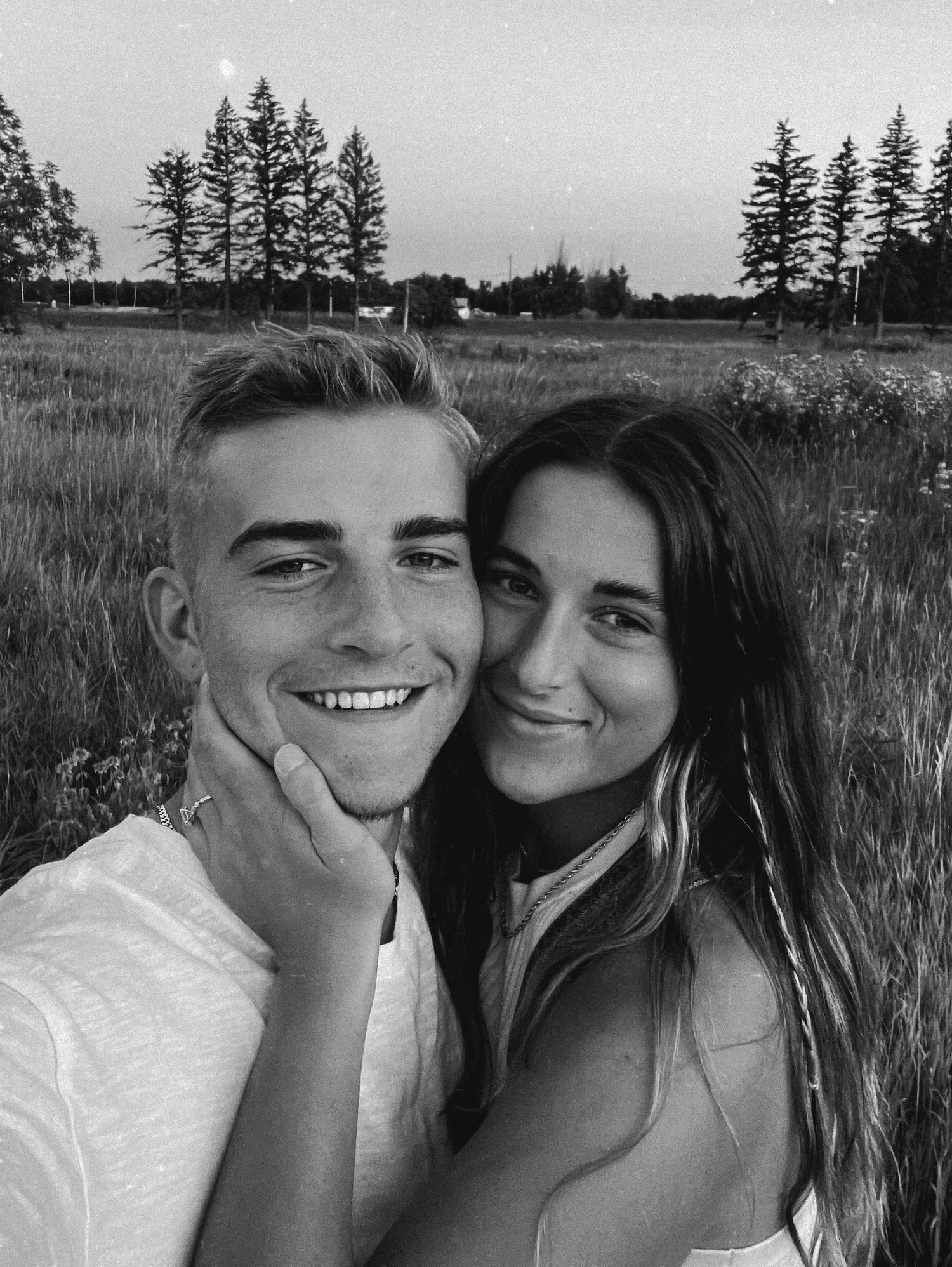 A couple shares intimate black and white moments together in a grassy field with evergreen trees in the background.