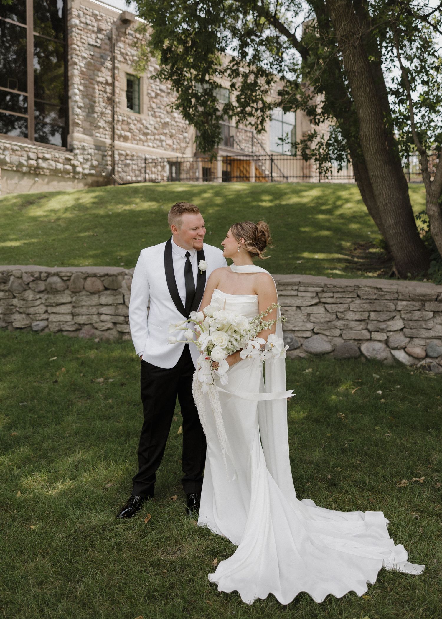 A couple in wedding attire shares a romantic moment together on a stone-walled garden lawn.
