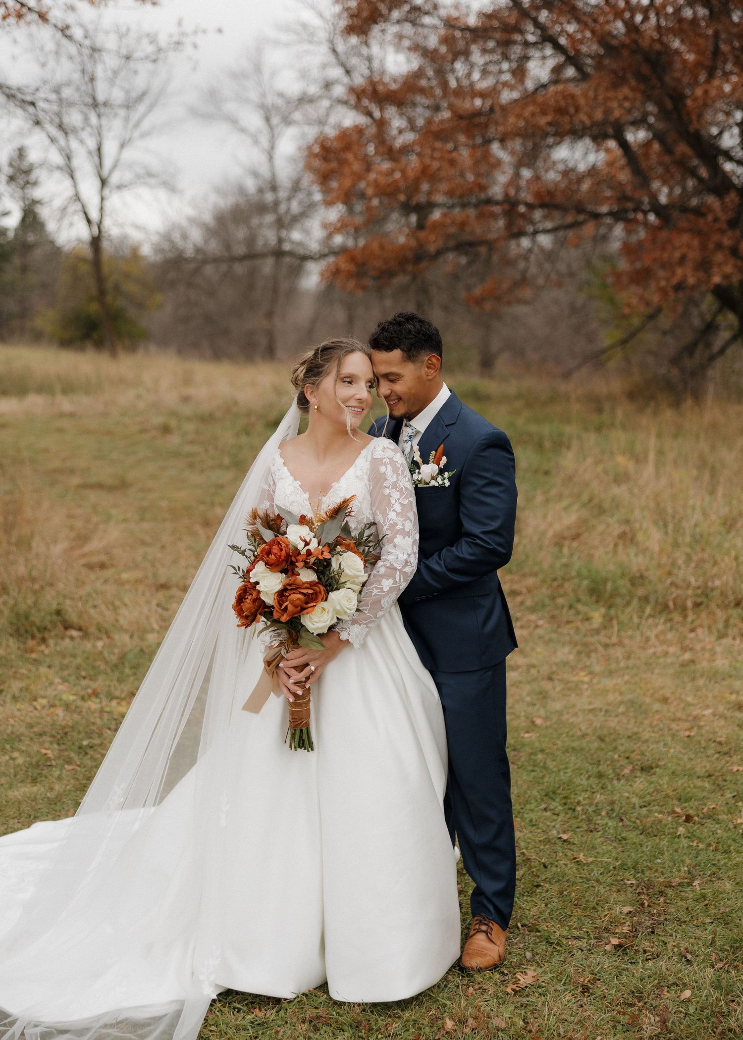 A couple in wedding attire share an intimate moment in a rustic autumn field with warm fall foliage in the background.