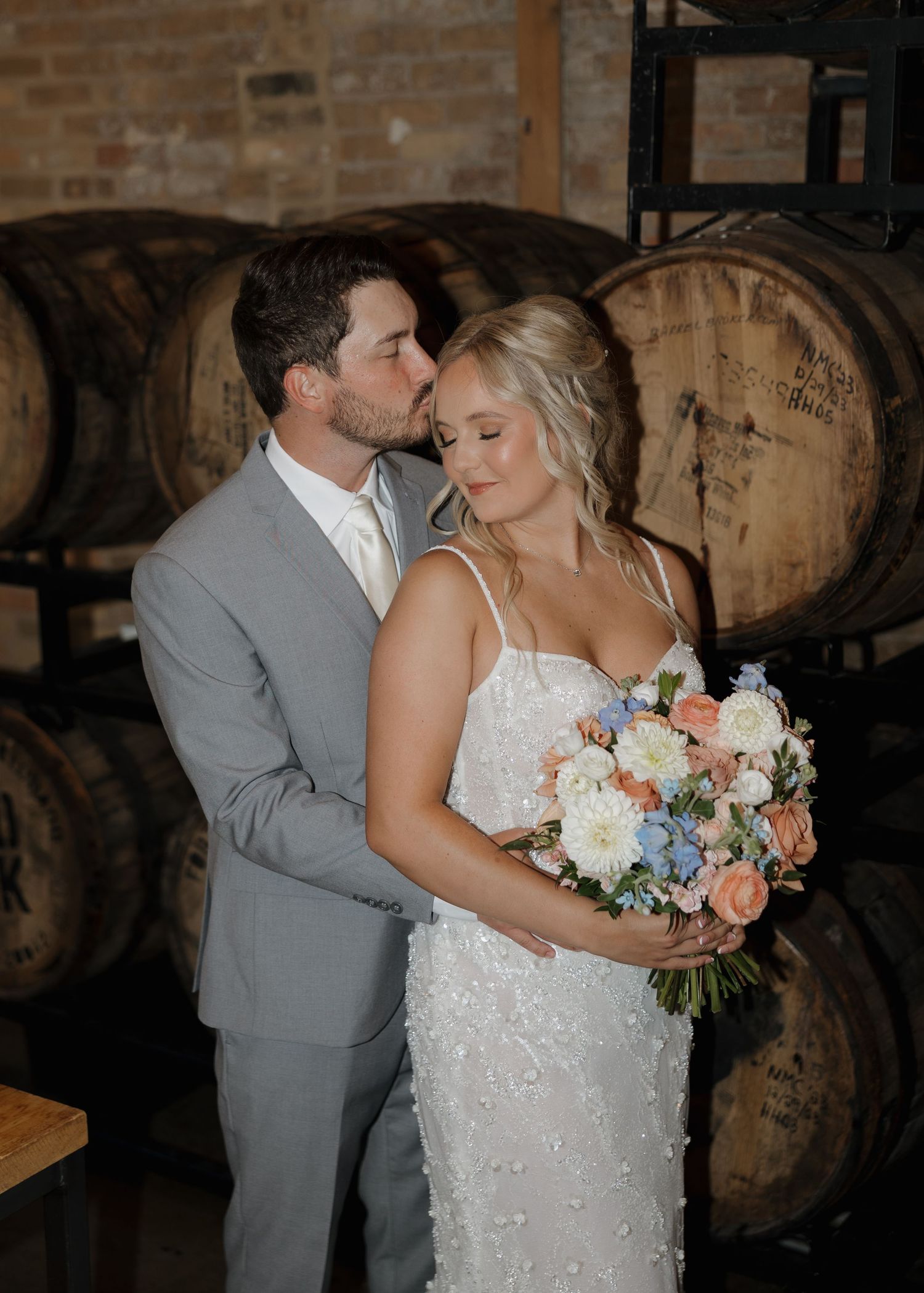 A couple shares an intimate kiss in a rustic barrel room setting with a beautiful bridal bouquet of peach and white flowers.