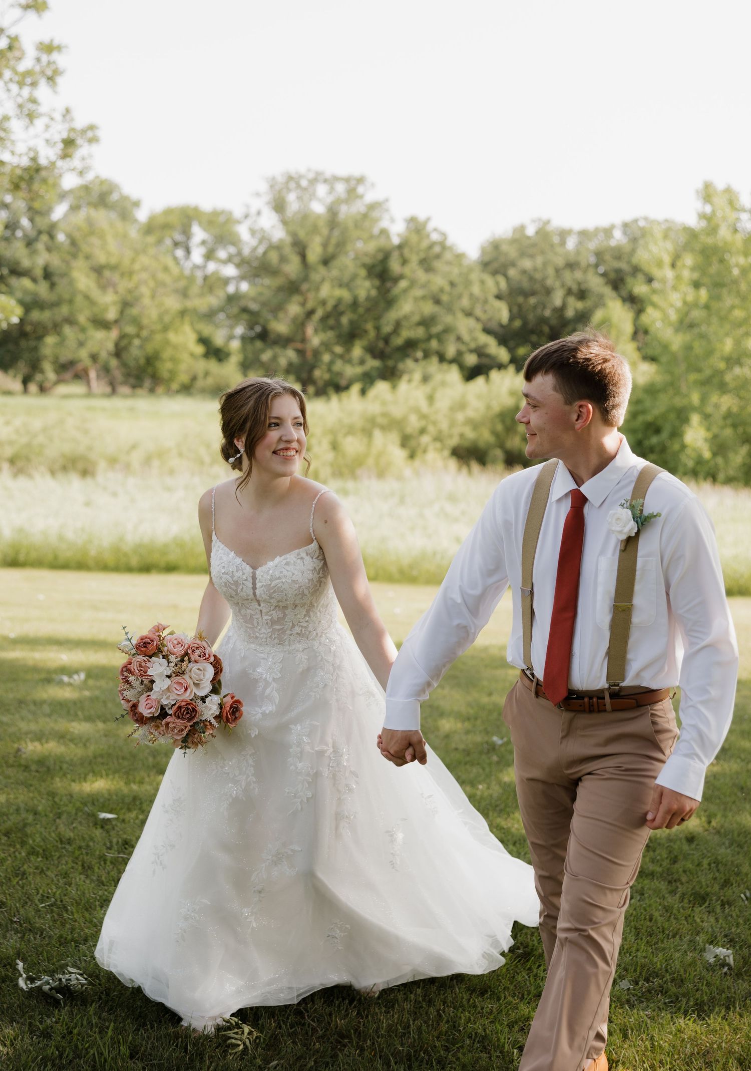 A couple in wedding attire dances playfully on a grassy lawn with trees in the background.