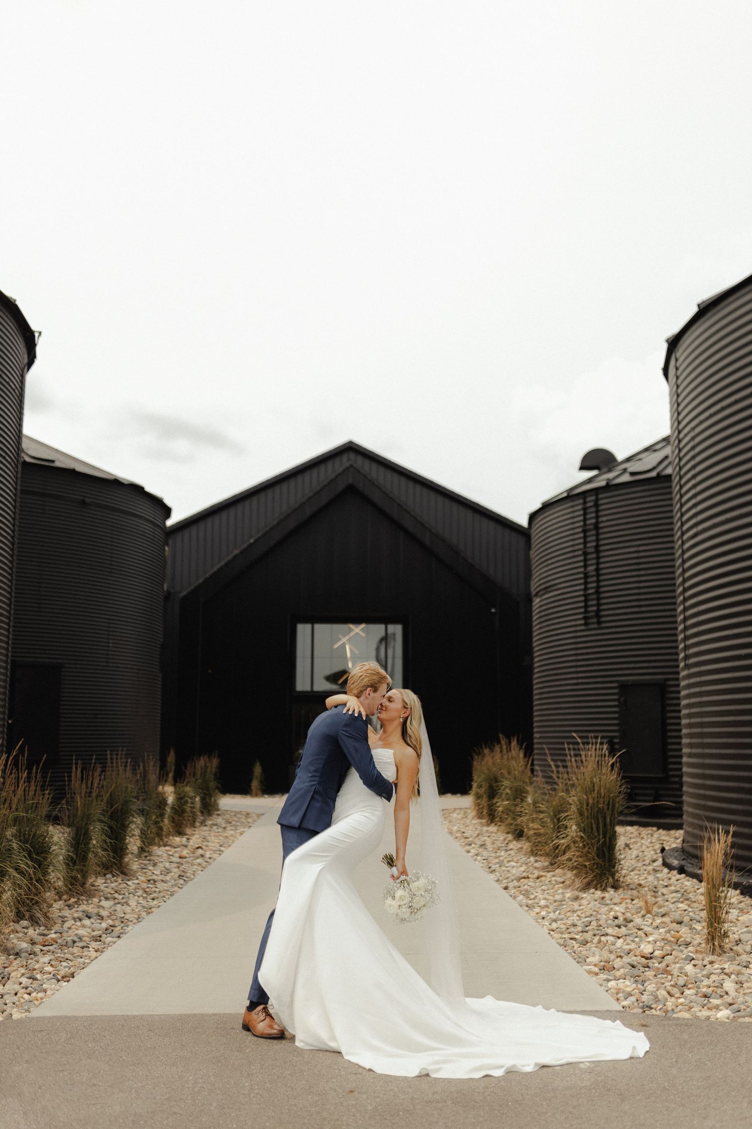 A romantic wedding photo sequence showing a couple in formal attire embracing against black beach huts on a pebble beach.