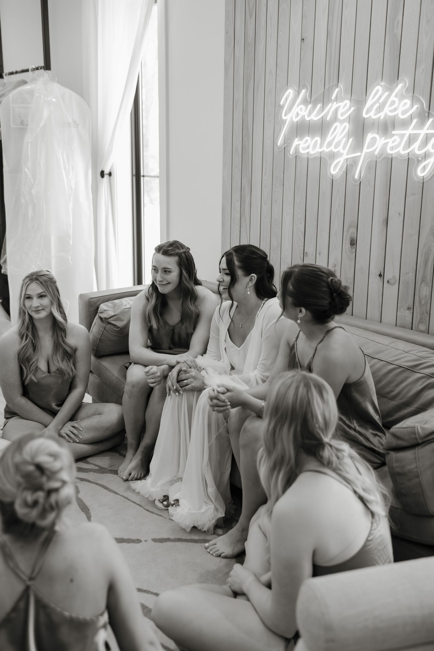 A group of women sit together on a couch under a neon sign that reads 'You're like really pretty' in white light.