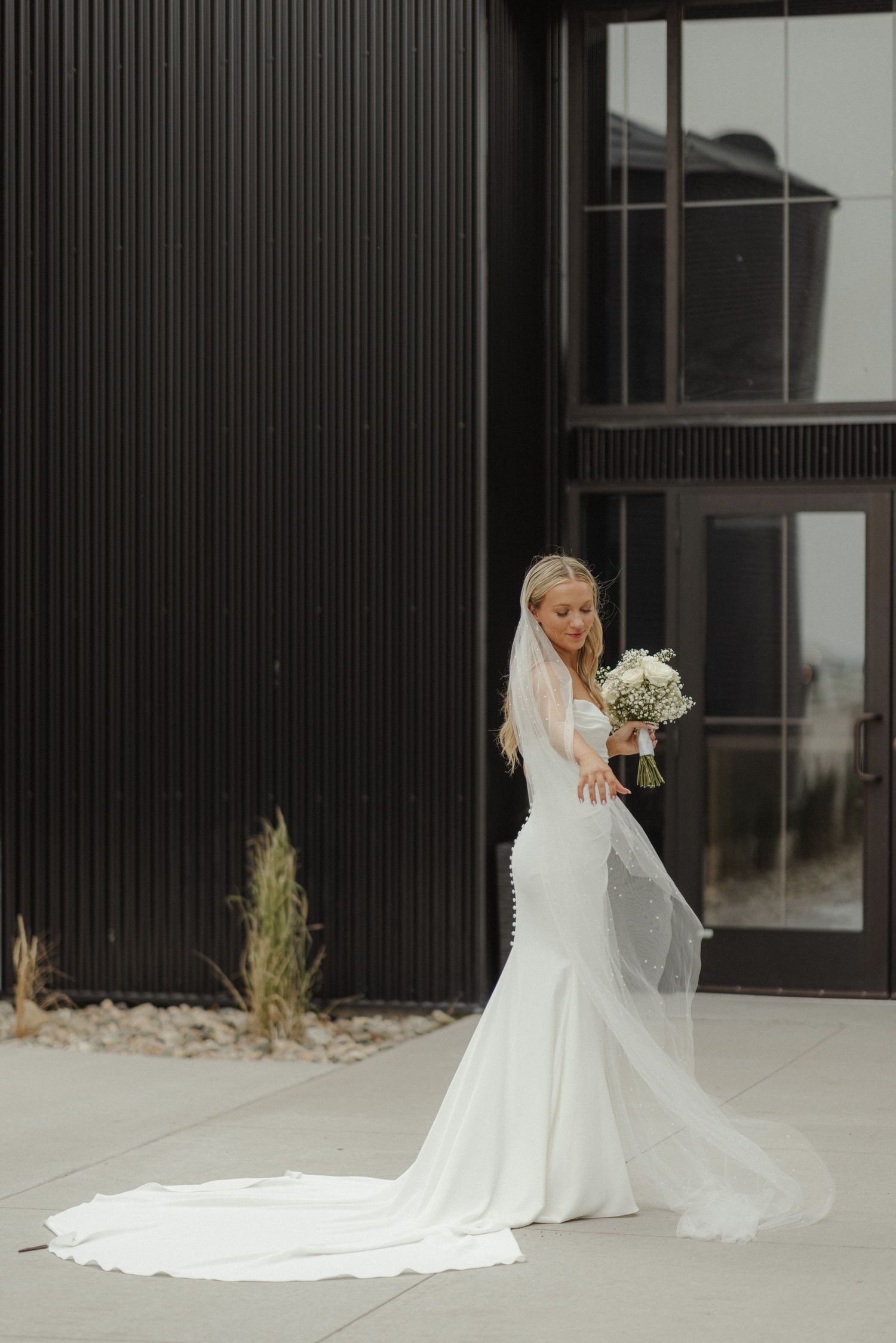 A stunning bride in a white wedding gown with a long cathedral veil poses elegantly against a modern black building.
