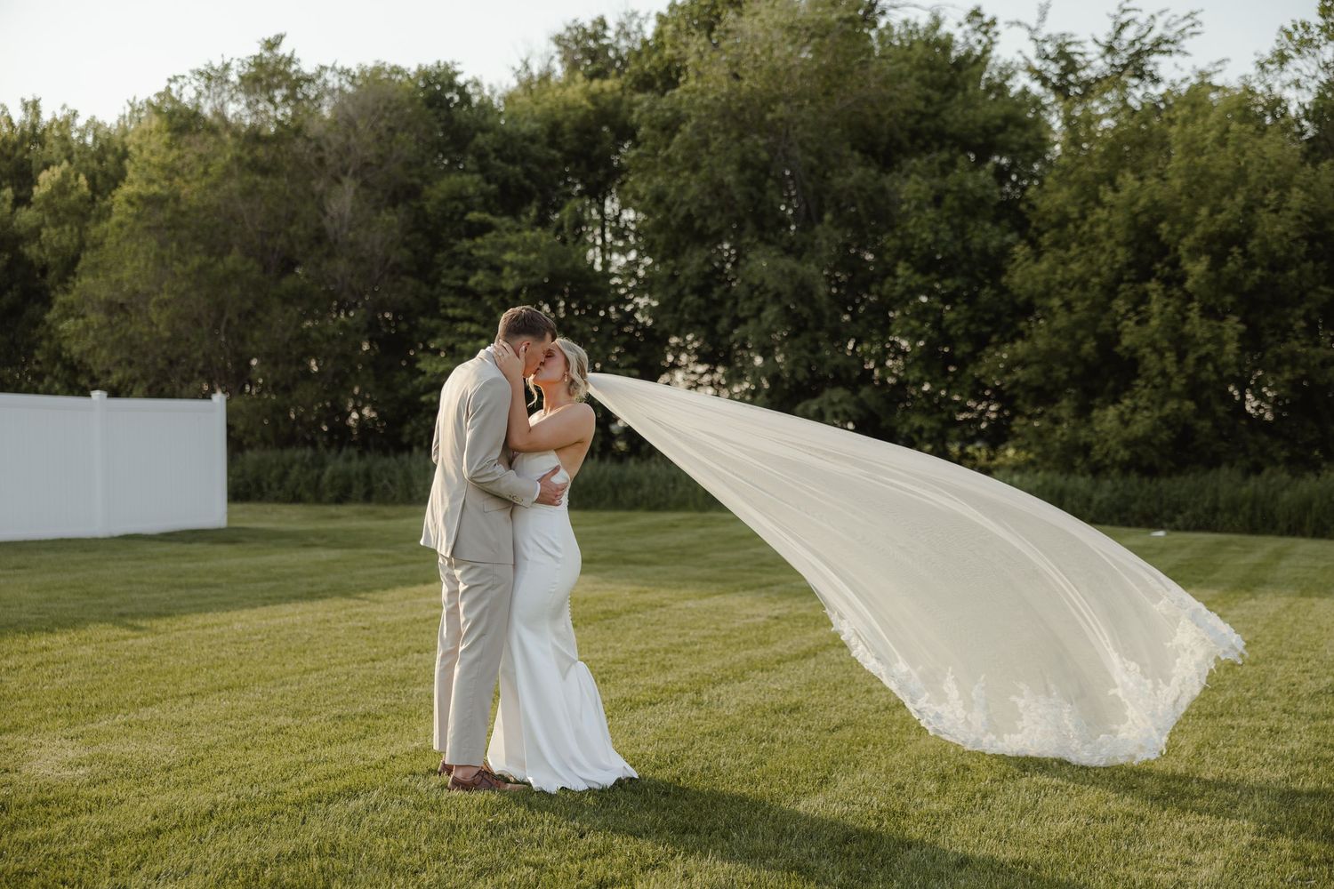 A couple stands together in a grassy field as a white wedding veil flows dramatically in the breeze.