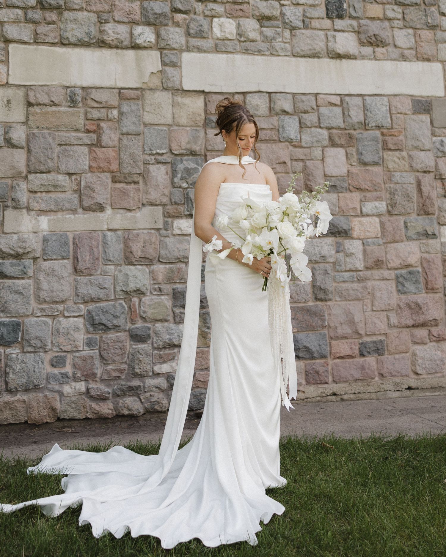 A bride in a flowing white strapless wedding gown poses against a rustic stone wall backdrop.