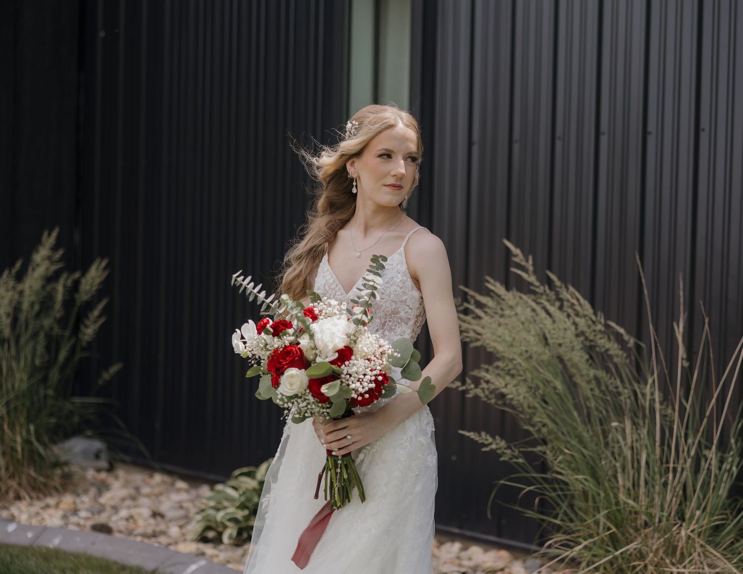 Bride in elegant white dress holds vibrant red and white bouquet while standing among ornamental grasses outdoors.