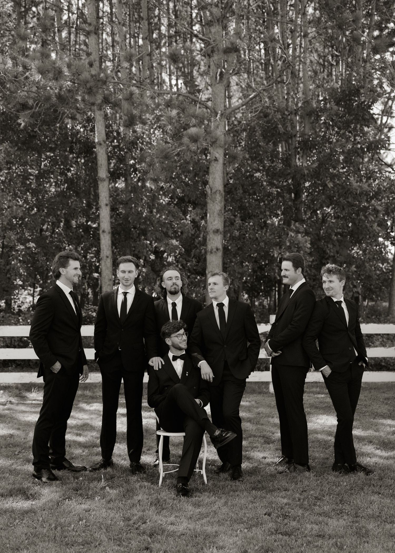 A group of people in black suits stand in a row outdoors against a backdrop of tall trees in a candid progression.