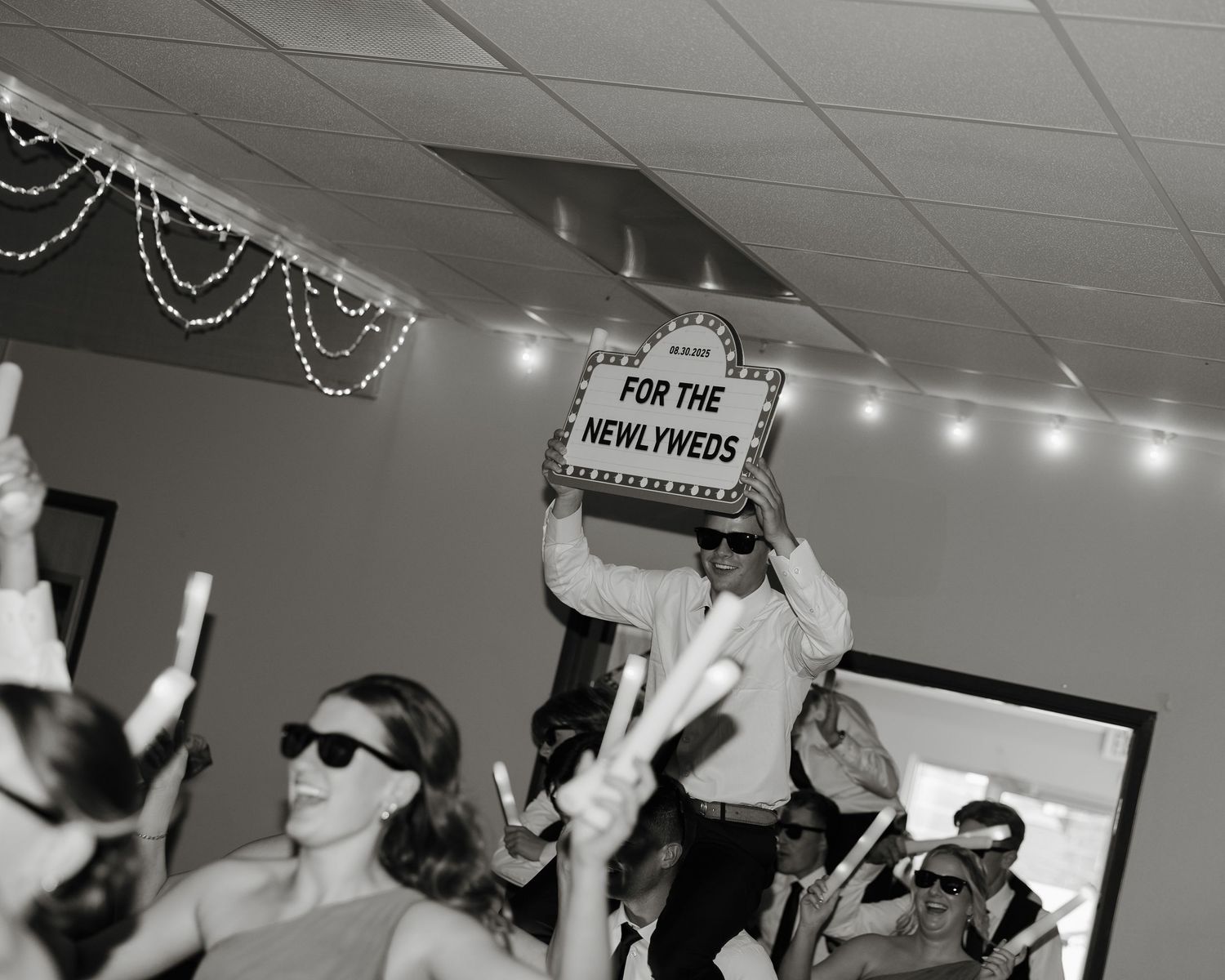 Wedding guests dance and celebrate on a crowded dance floor at a reception with string lights overhead.