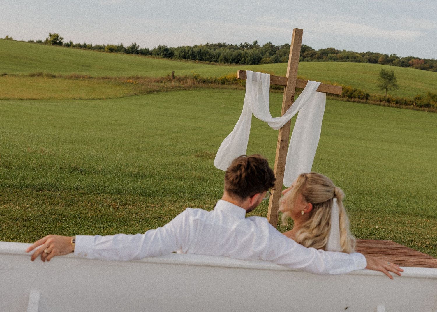 A couple sits together on a blanket in a grassy field with a wooden cross and white fabric in the background.