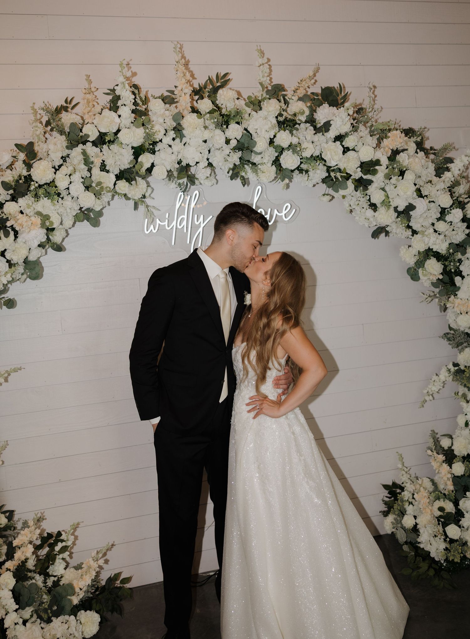 Romantic wedding scene with couple kissing under an elegant white floral arch with neon 'happily ever after' sign.