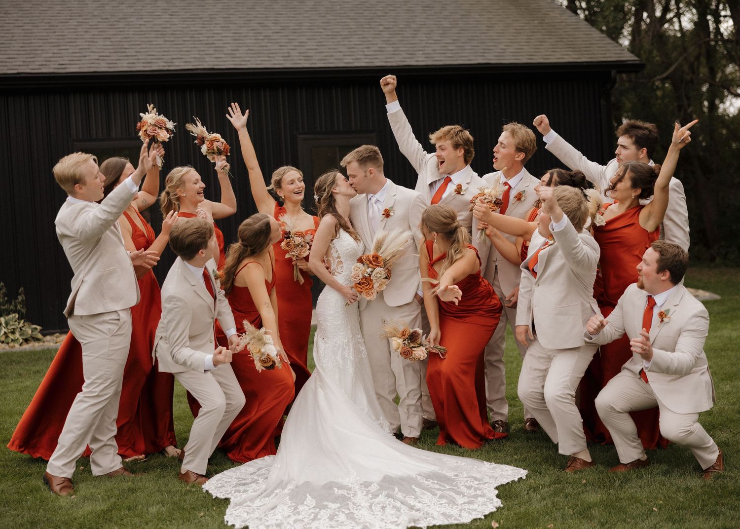 Wedding party in white and coral attire poses joyfully on grass during outdoor celebration.