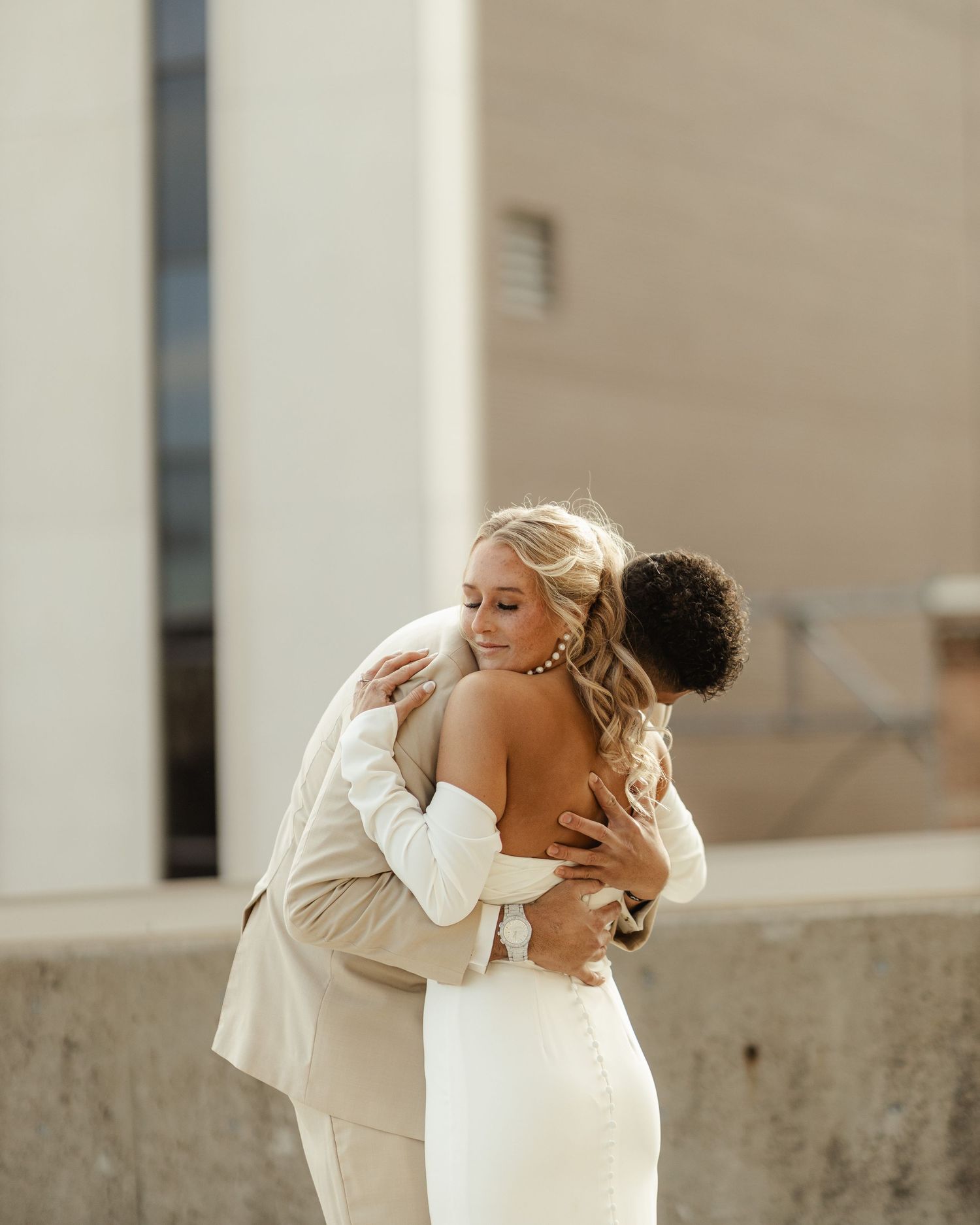 Two people in white outfits embrace tenderly while sitting on concrete steps in warm sunlight.