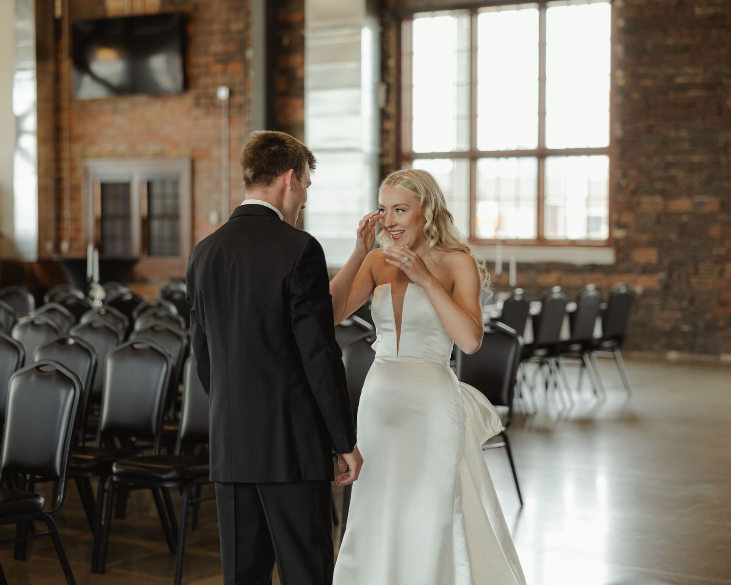 A bride and groom share an intimate moment in a rustic brick venue with large windows and natural light.