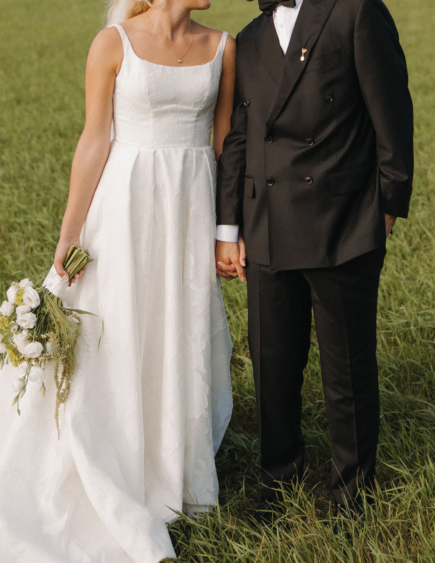 A bride in a white dress and groom in black suit hold hands while walking through a sunlit grassy field.