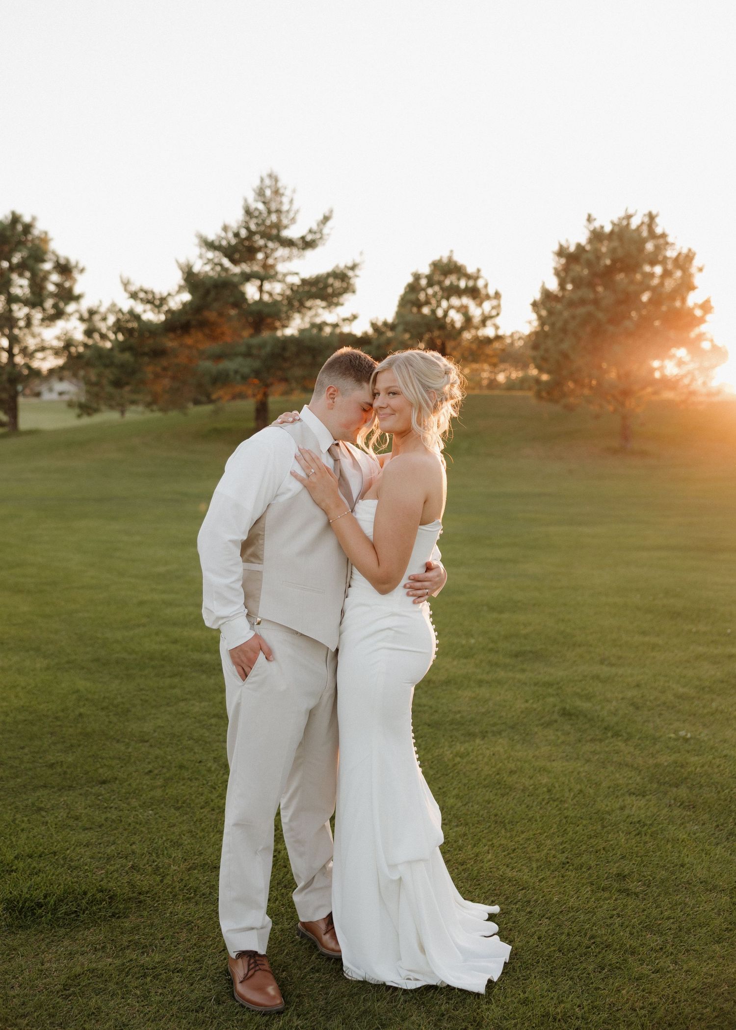 A romantic wedding couple shares tender moments on a grassy hillside during golden hour sunset.