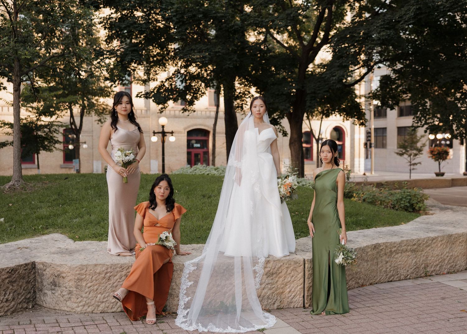 Wedding party members in coordinated dresses pose together outdoors on a stone pathway near historic buildings.