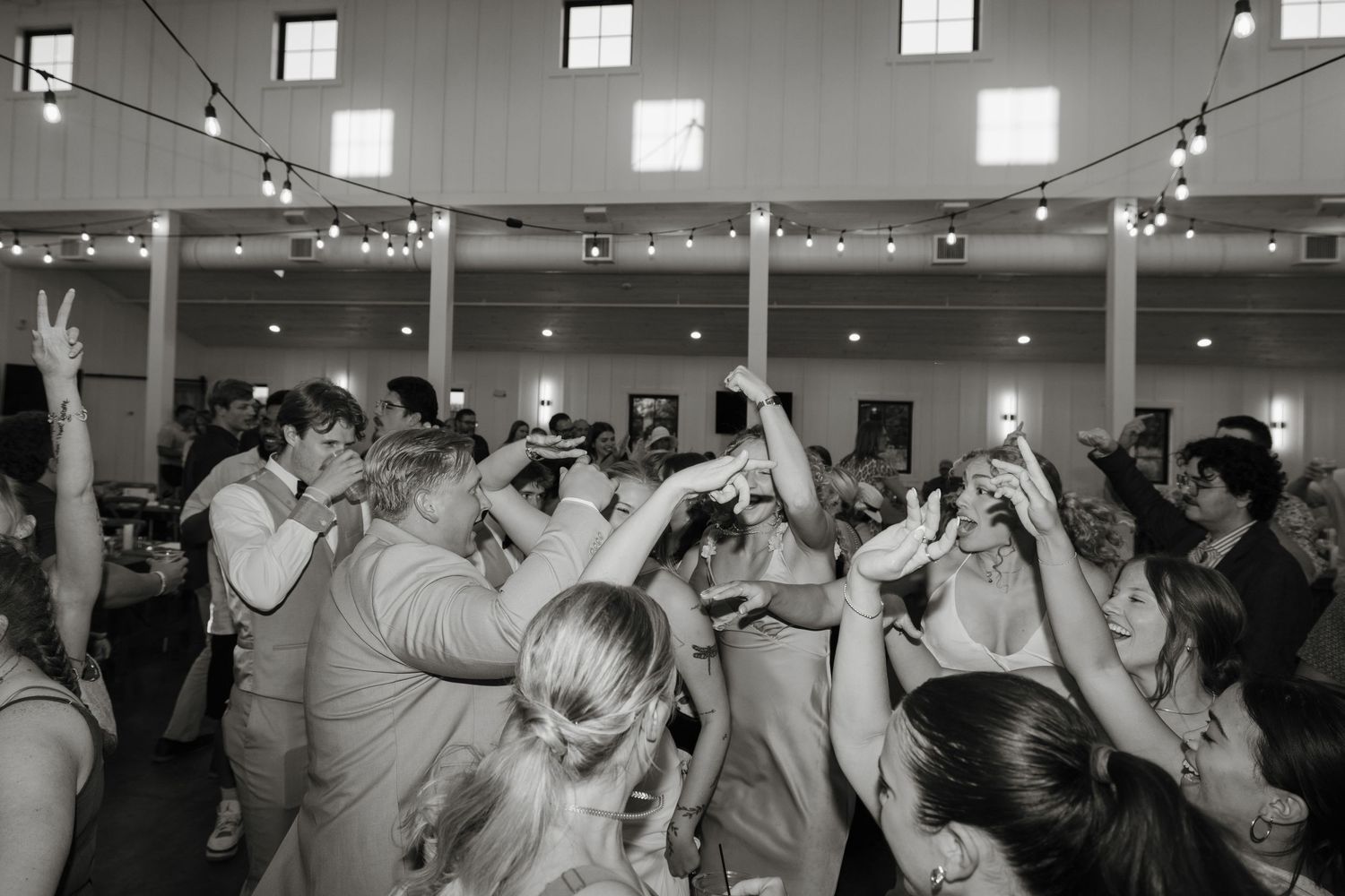 A black and white photo of a crowded dance floor at a reception with people dancing with their hands in the air.