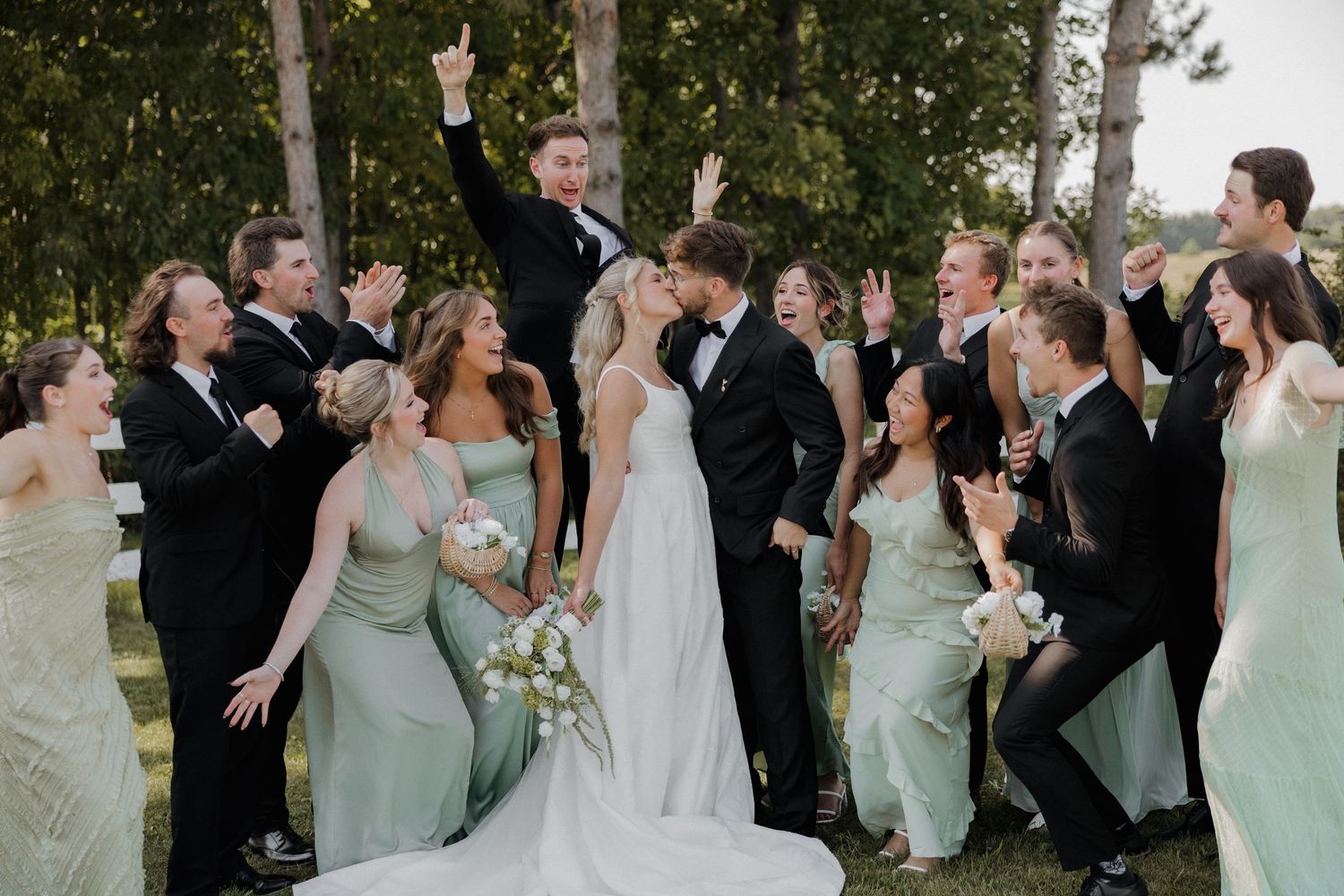 A wedding party in mint green bridesmaid dresses and black suits celebrates joyfully outdoors with raised arms.