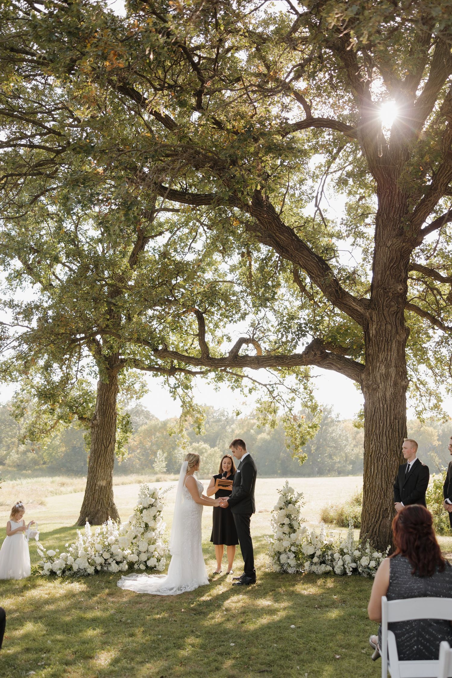 A romantic outdoor wedding ceremony takes place under majestic oak trees with sunlight streaming through the branches.