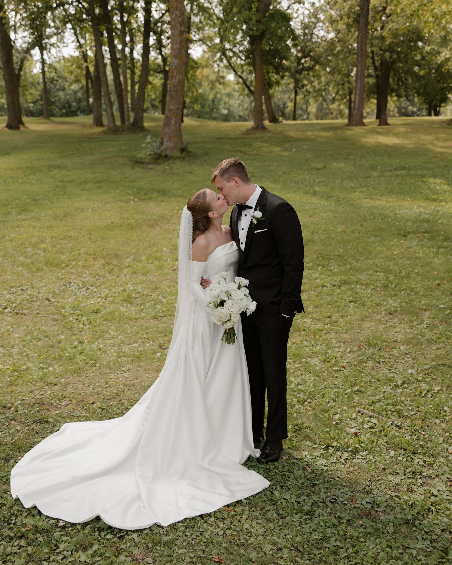 A couple shares an intimate moment on their wedding day in a serene park setting with tall trees and green grass.