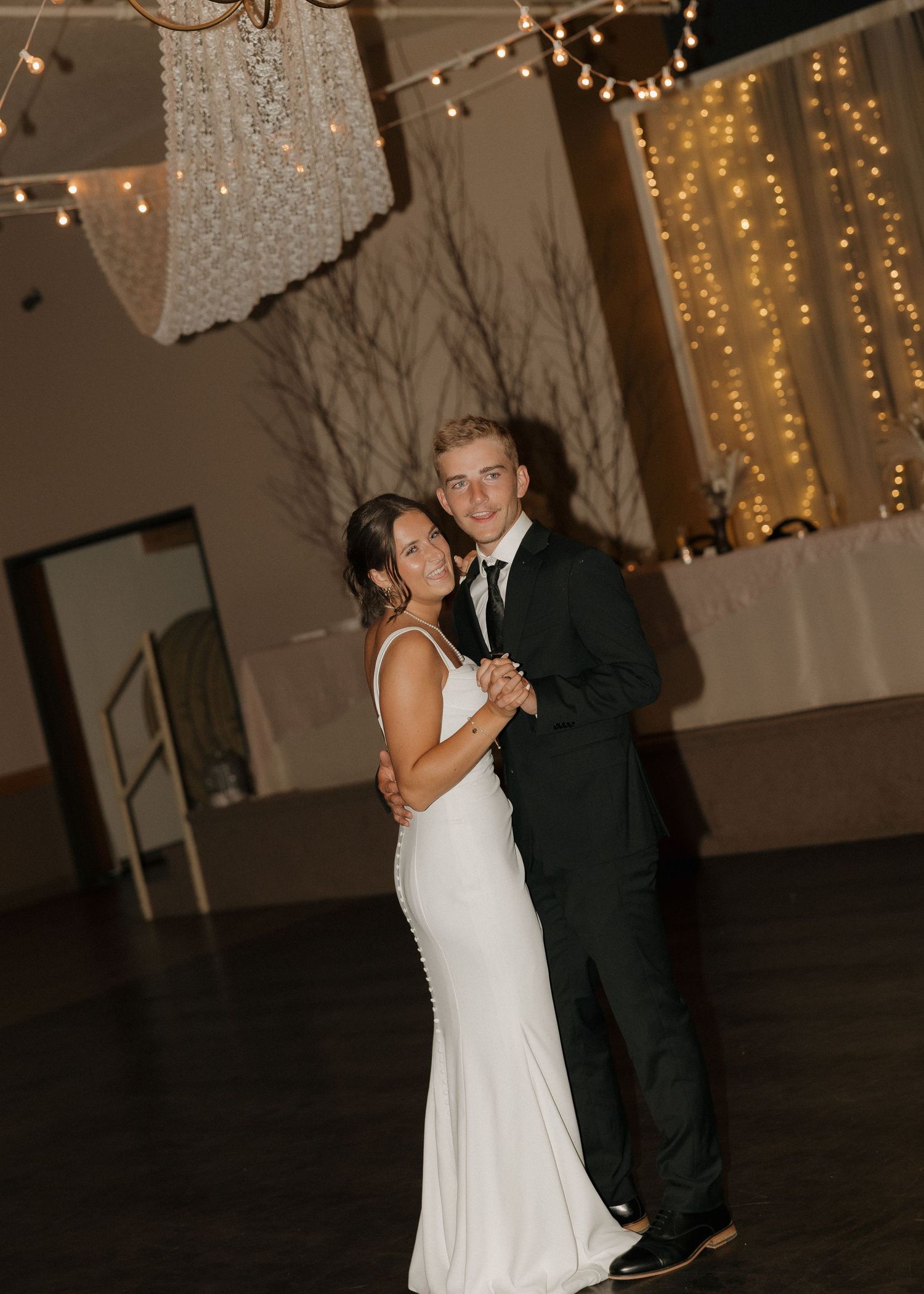 A couple shares their first dance at a wedding reception with string lights and drapery in the background.