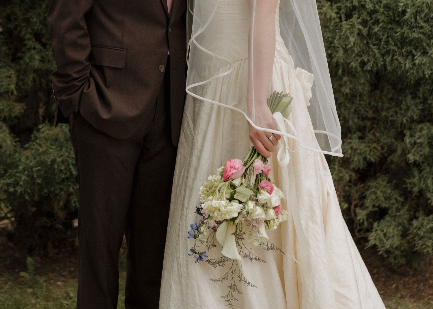 Bride in ivory gown holding pink and white bouquet stands beside groom in dark suit outdoors.