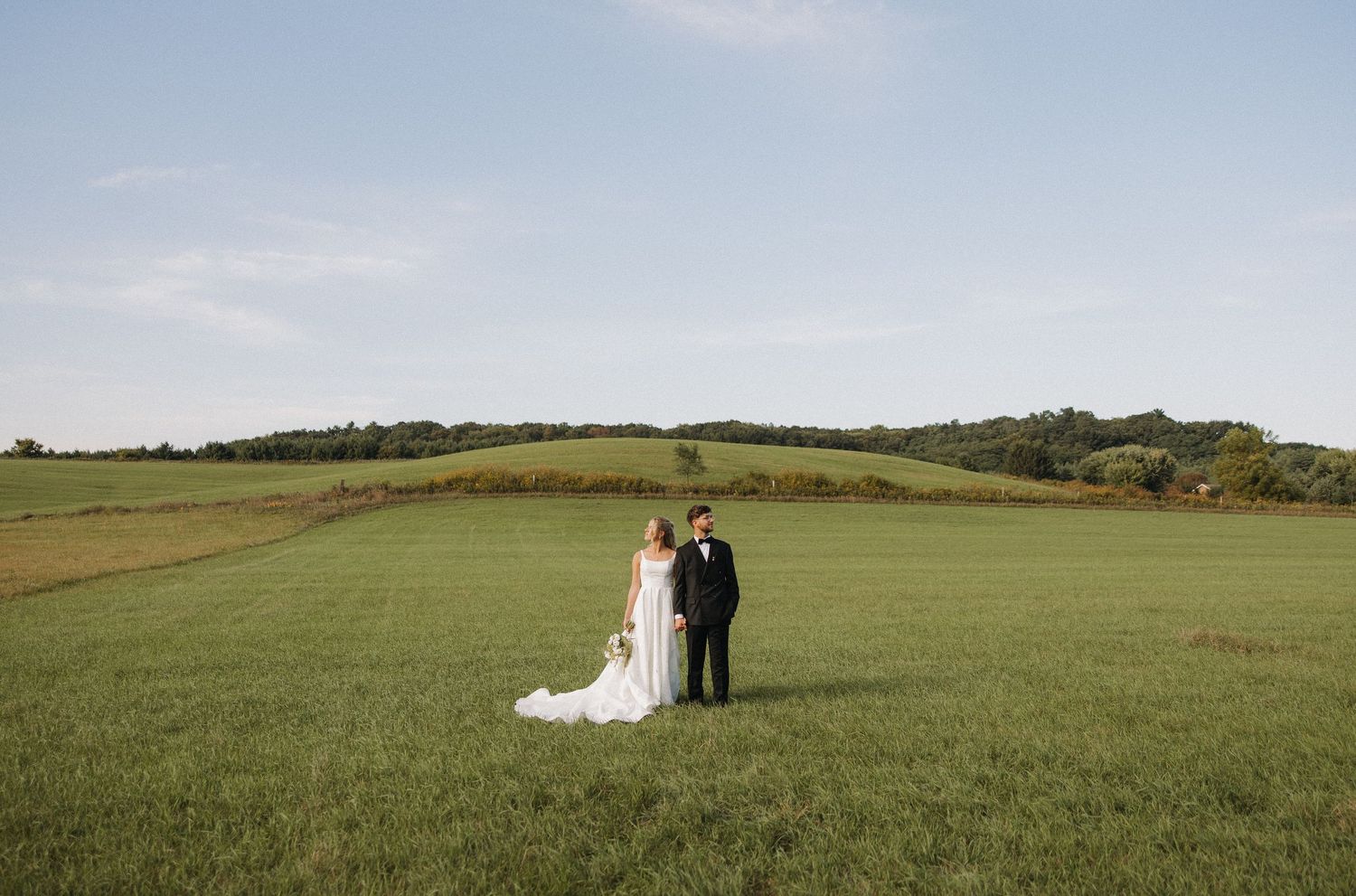 A couple stands together in a vast green meadow with rolling hills and scenic countryside views in the background.